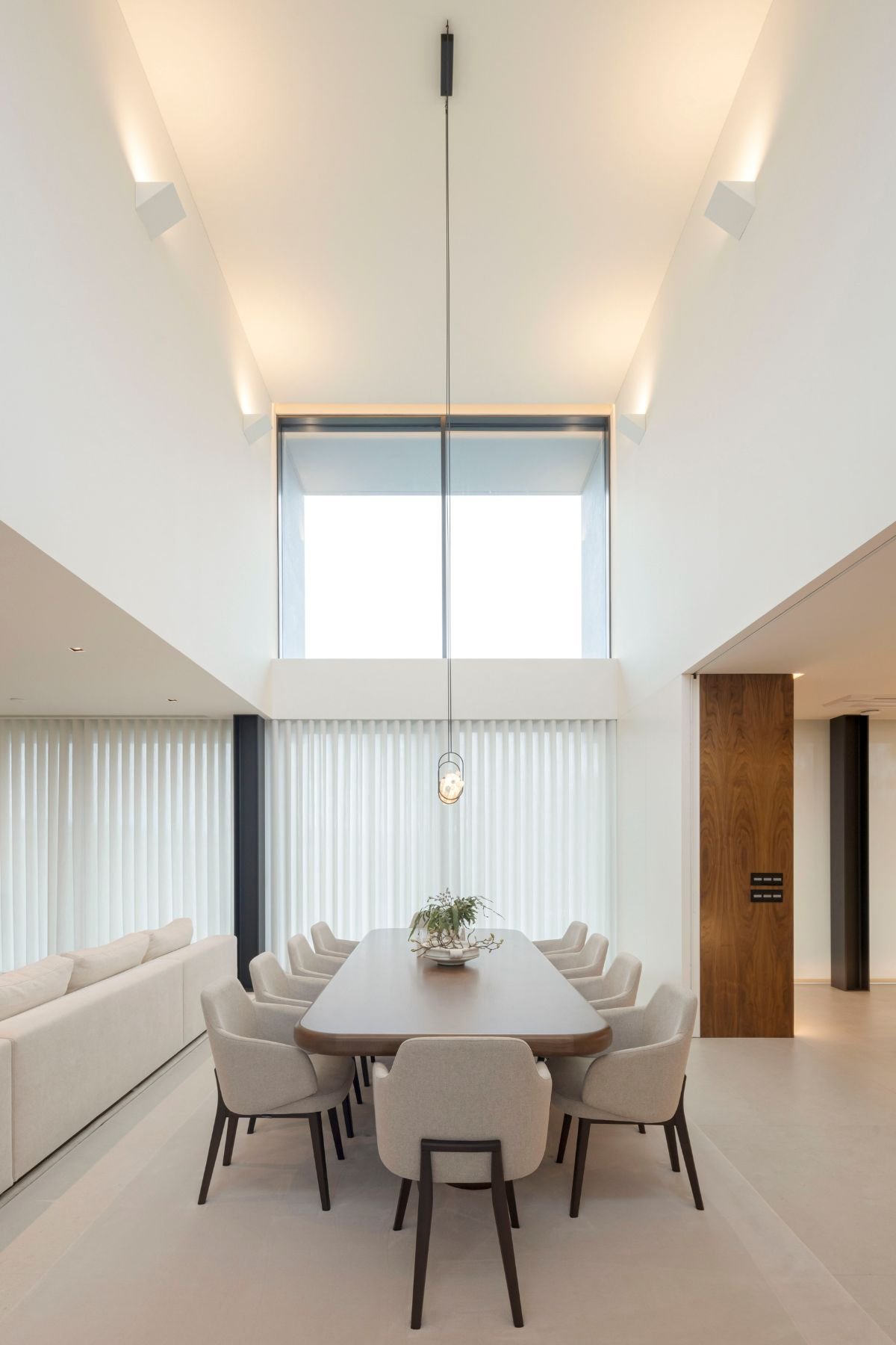 Sala de jantar com mesa de jantar retangular em madeira, cadeiras de jantar beges com pernas em madeira e pé-direito duplo luminoso.