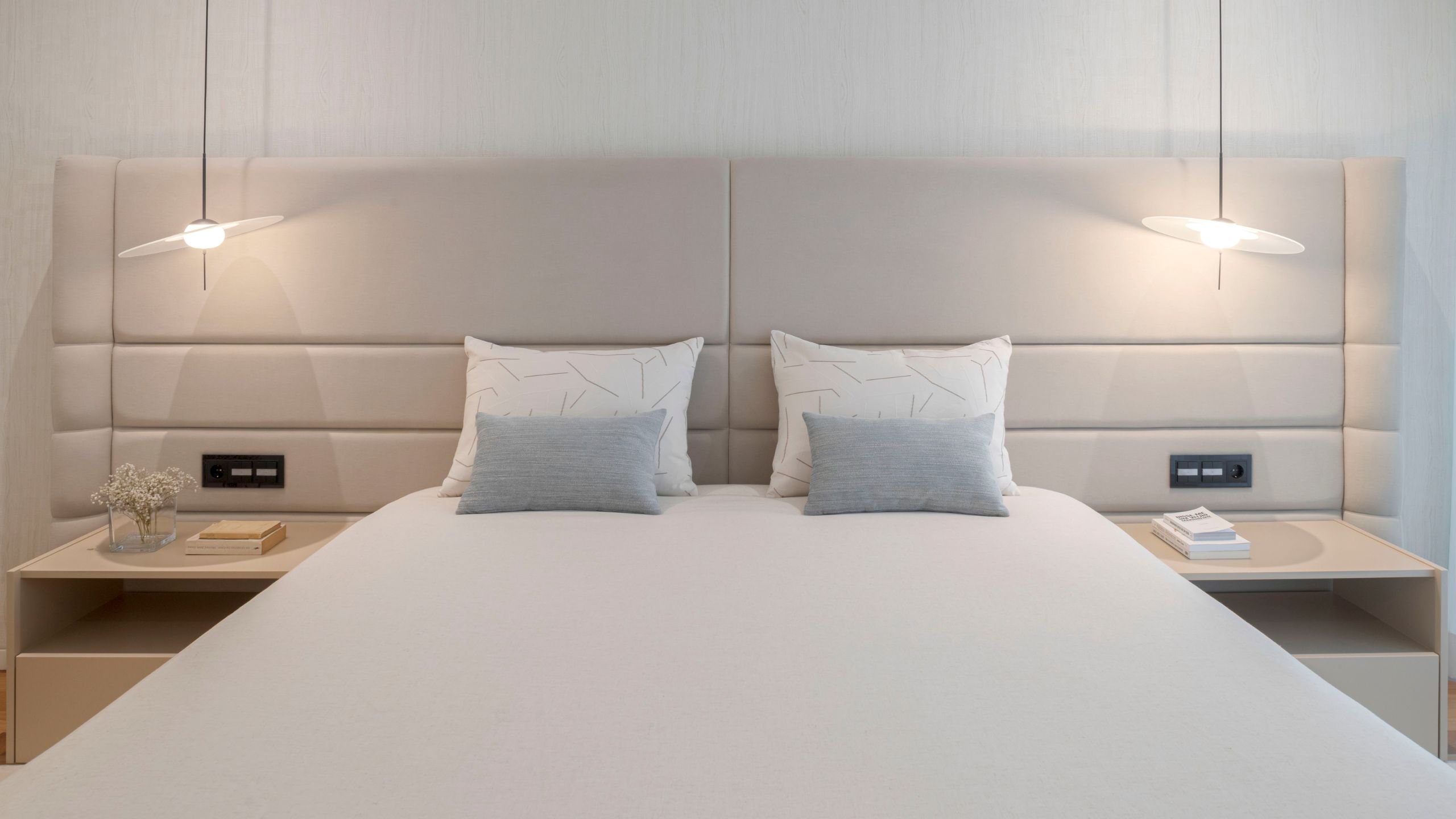 Cama de casal com cabeceira de cama estofada em tom neutro, mesas de cabeceira integradas e candeeiros de suspensão numa suite principal.