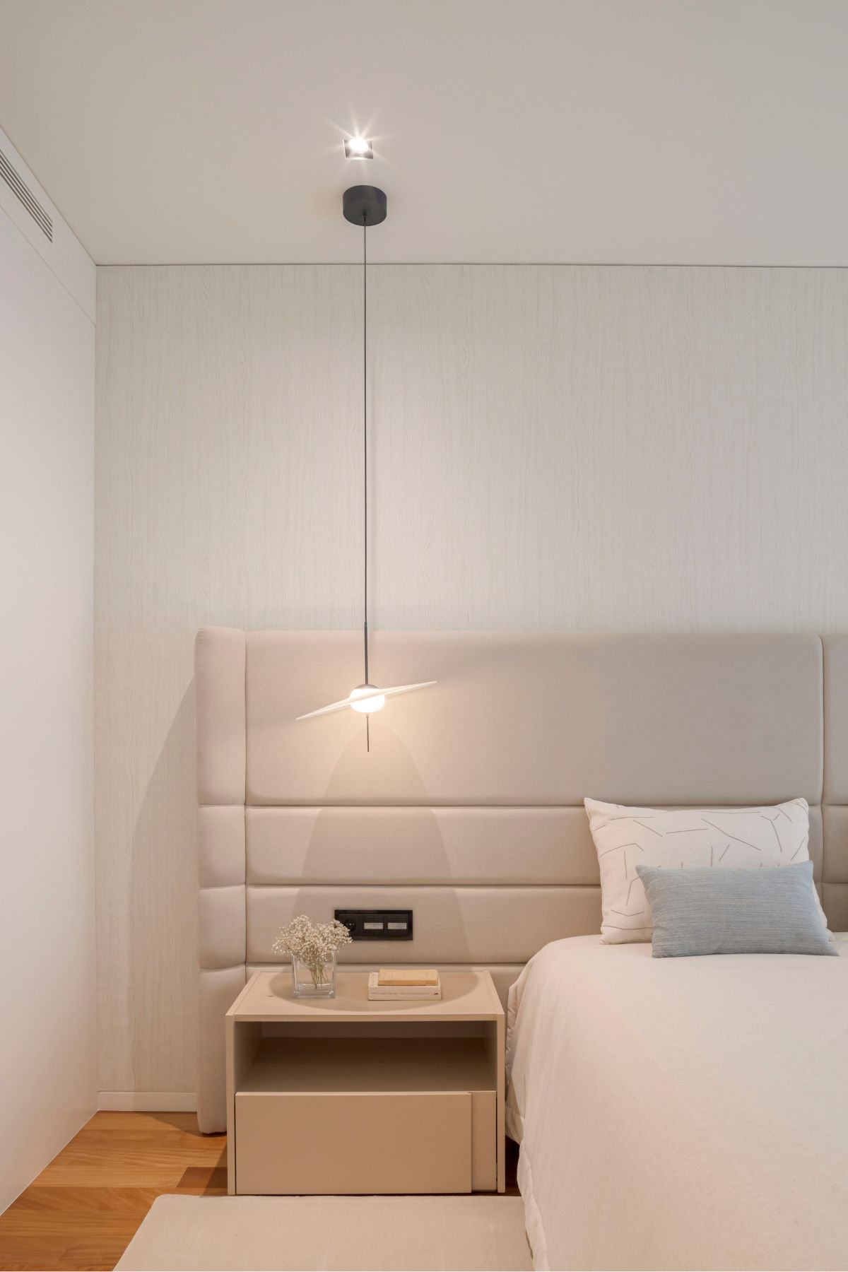 Pormenor da mesa de cabeceira bege com jarra decorativa e candeeiro de suspensão moderno suspenso junto à cabeceira de cama estofada.