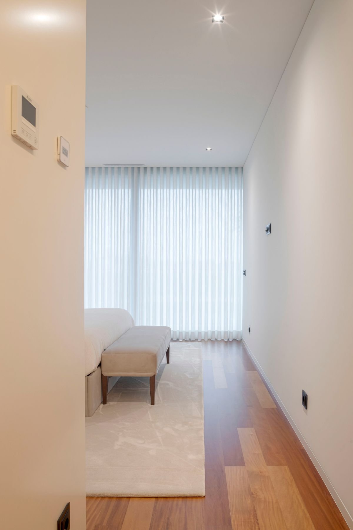 Perspetiva da entrada da suite principal mostrando o banco de apoio aos pés da cama de casal junto à grande janela com cortinados.