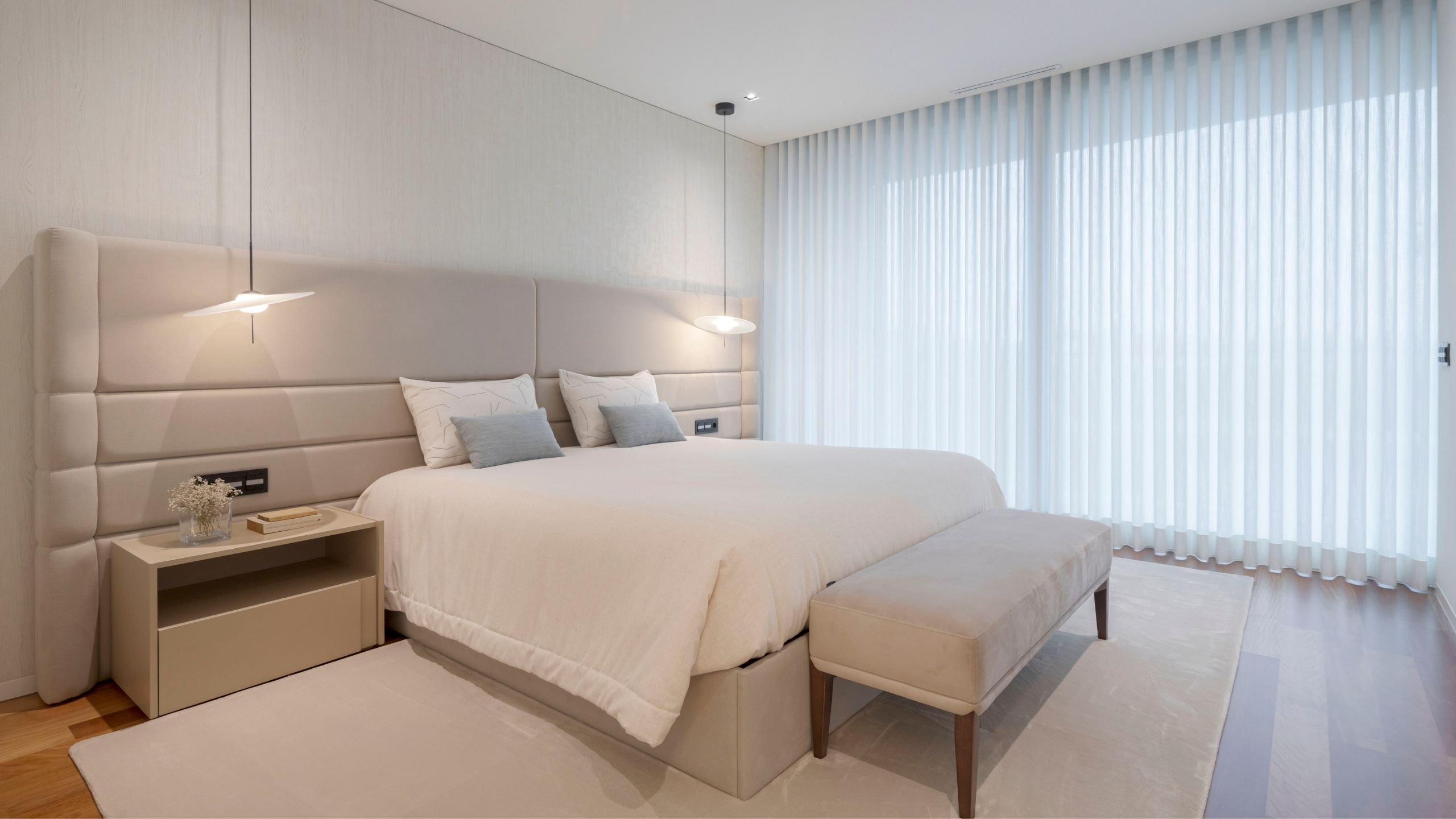 Vista geral do quarto de casal moderno com cama de casal, banco de apoio, mesas de cabeceira e cortinas brancas translúcidas.