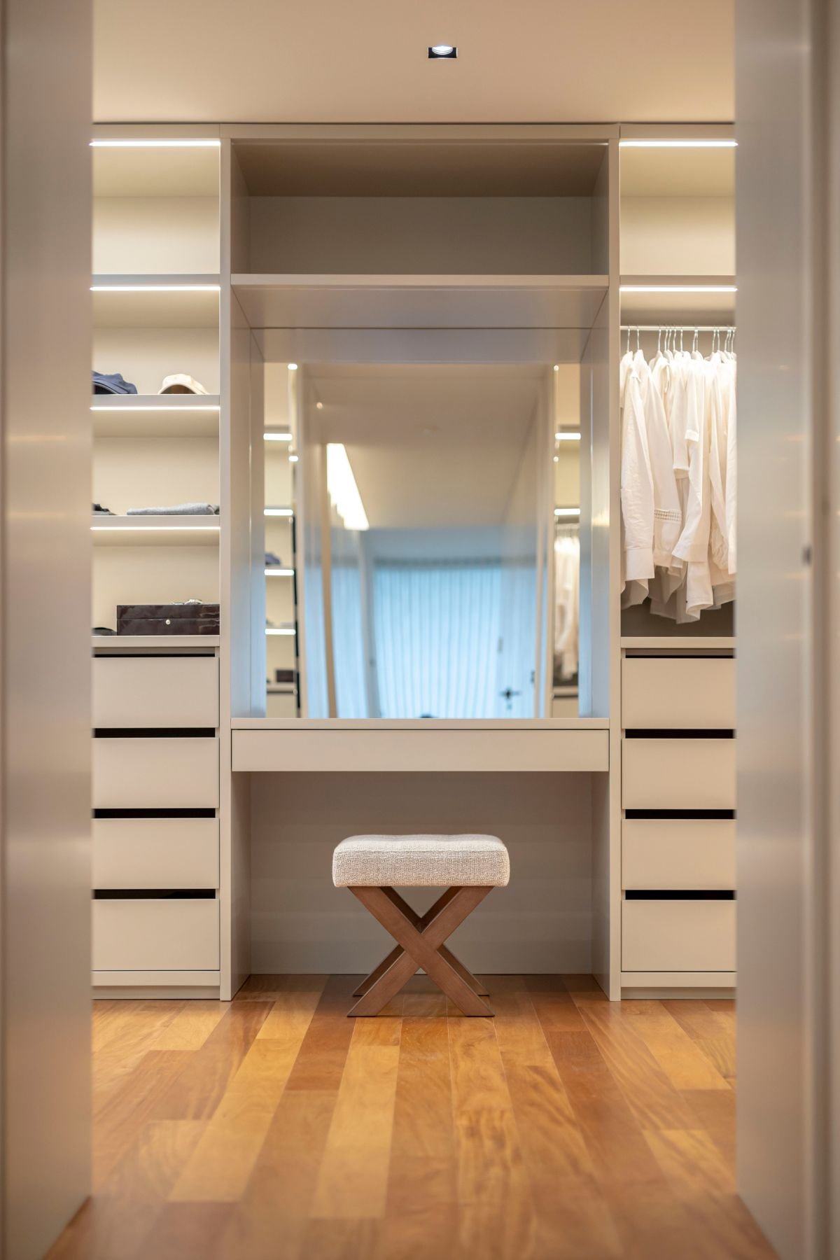 Walk-in closet por medida com armários iluminados, toucador central com espelho e banco de apoio com pernas em X.