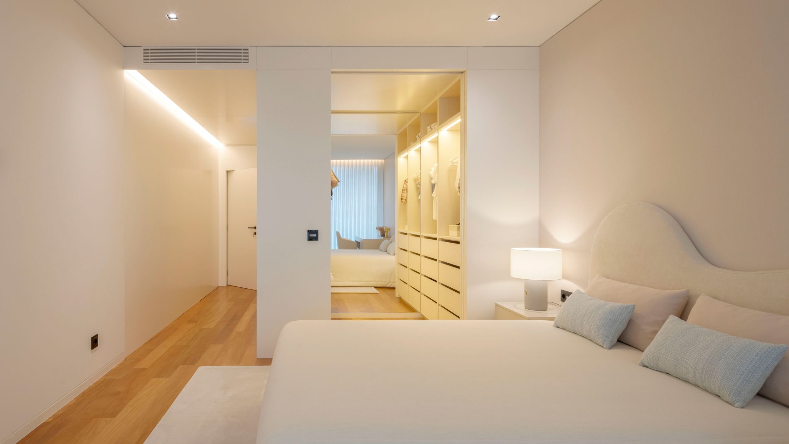 Quarto juvenil luminoso com cama de casal, cabeceira de cama ondulada, mesa de cabeceira e closet aberto ao fundo.