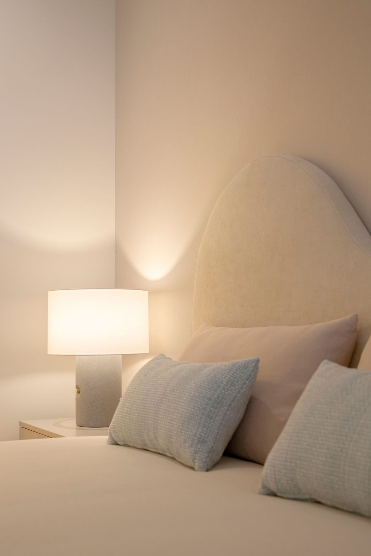 Pormenor da cabeceira de cama estofada em tom neutro com linhas orgânicas e candeeiro de mesa em quarto juvenil.