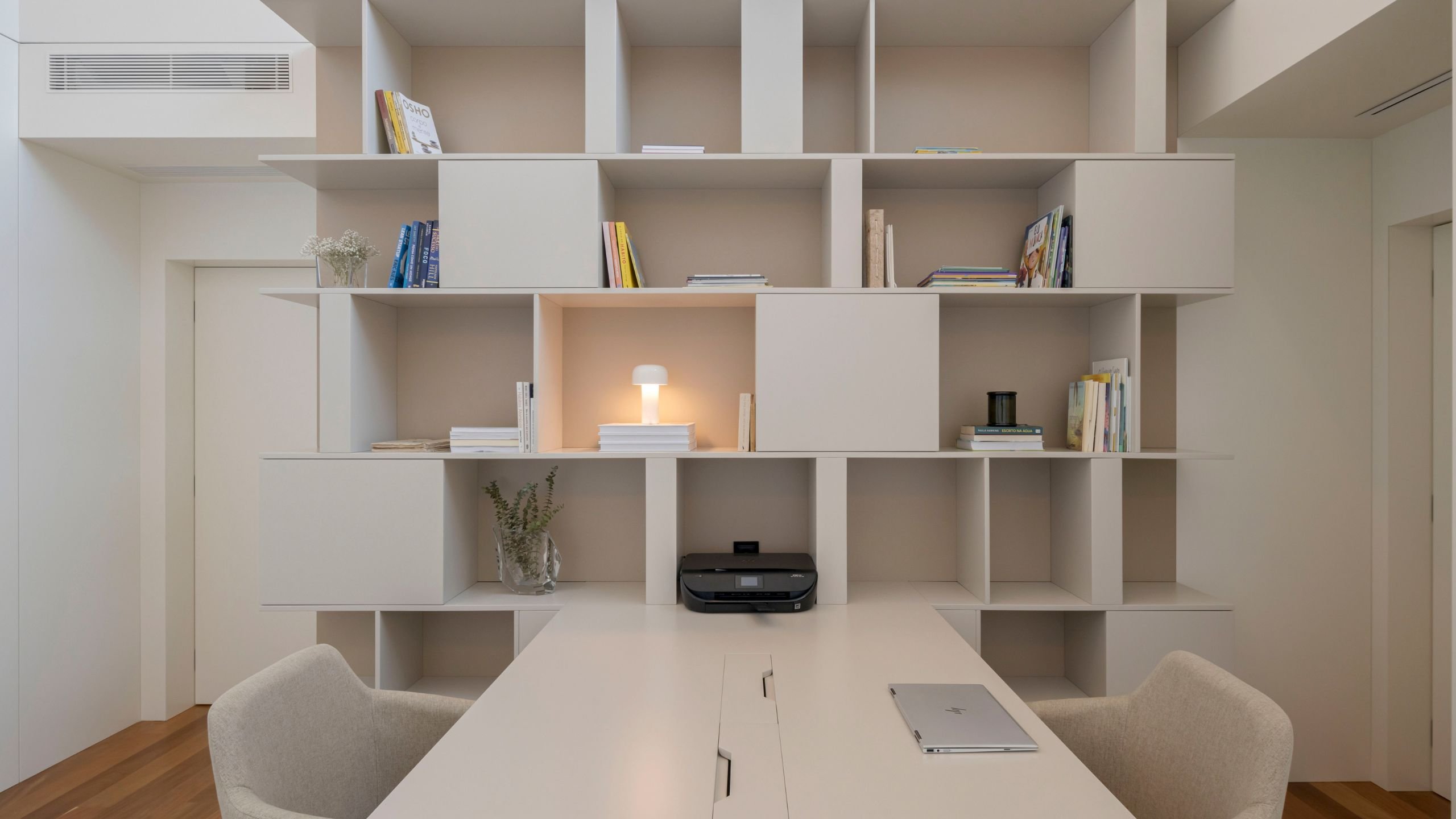 Escritório moderno com estante de livros modular, secretária dupla e cadeiras de escritório estofadas em tom neutro.