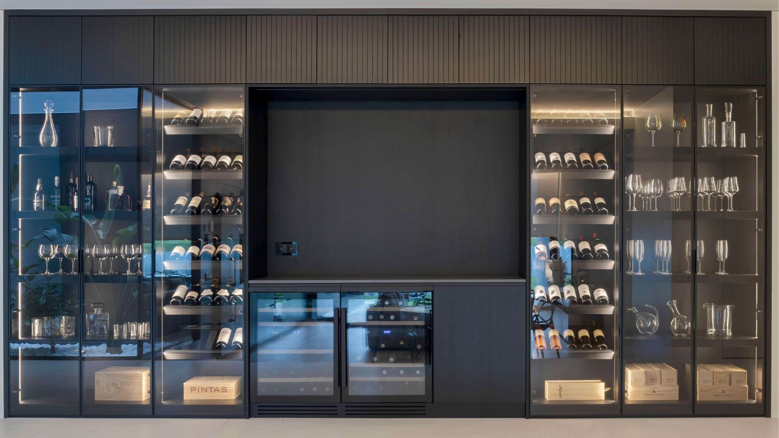 Garrafeira moderna por medida com armários pretos, vitrines de vidro iluminadas para vinhos e copos, e garrafeiras climatizadas.