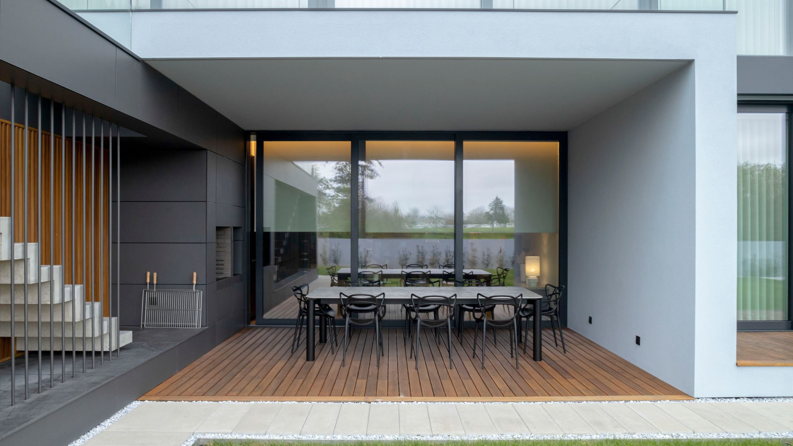 Alpendre moderno de moradia com mesa de jantar exterior em pedra terrazzo cinza, cadeiras de design pretas e deck de madeira natural, ao lado de churrasqueira encastrada e escada flutuante.