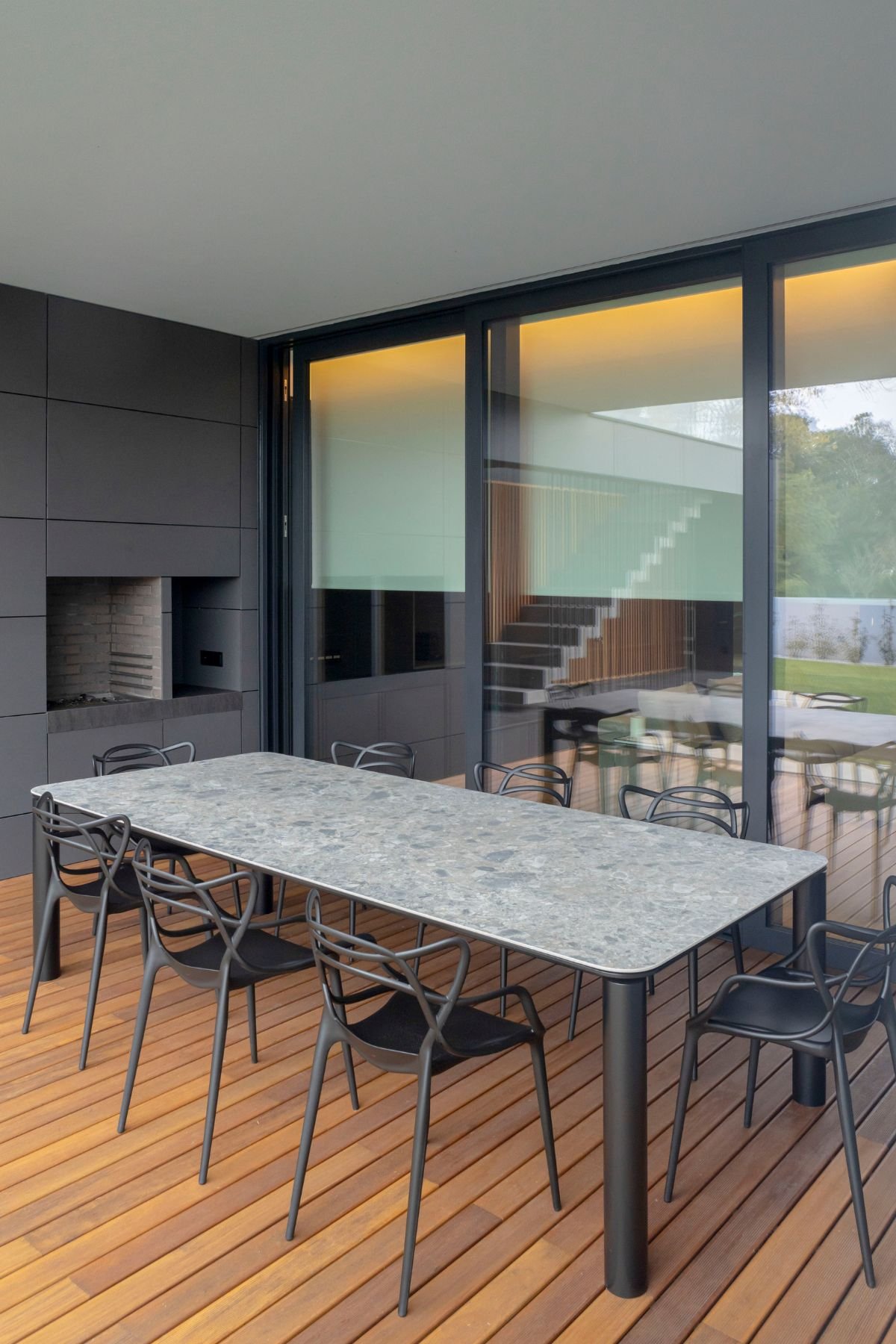 Mesa de jantar por medida com tampo em pedra cinzenta e pernas pretas, cercada por cadeiras de exterior pretas sob alpendre com deck de madeira e zona de churrasqueira.