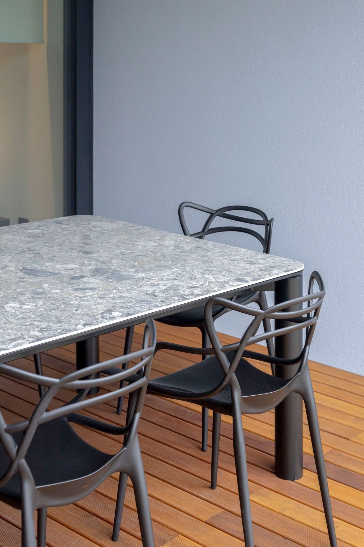 Detalhe do tampo em pedra estilo terrazzo de mesa de jantar de exterior com cadeiras pretas de design e deck de madeira em zona de refeições ao ar livre.