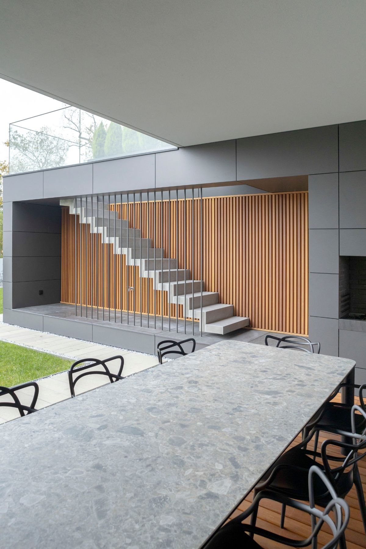 Perspetiva de mesa de jantar exterior em pedra cinza e cadeiras pretas em alpendre contemporâneo, com escada flutuante de betão e painel de ripas de madeira ao fundo.