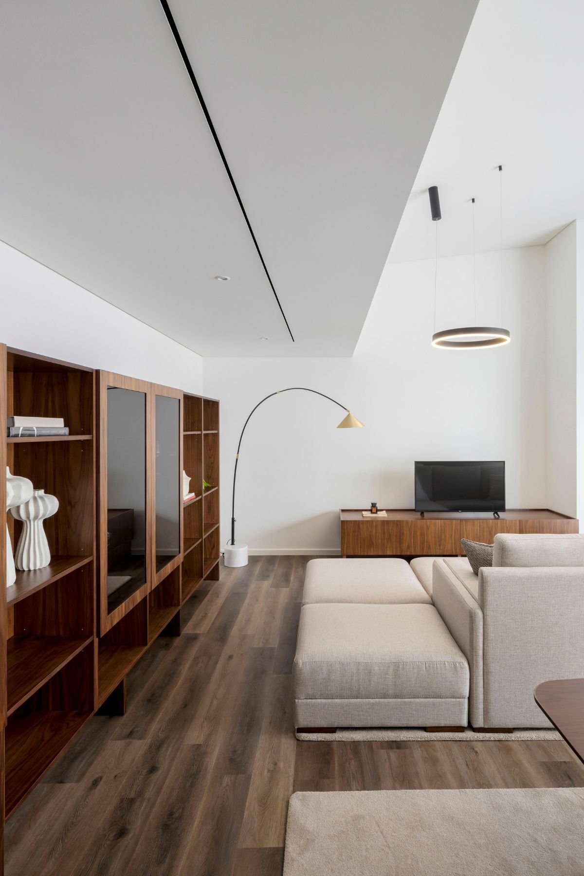 Vista lateral da zona de estar minimalista com móvel de tv em nogueira, candeeiro de pé curvo e estante de livros.