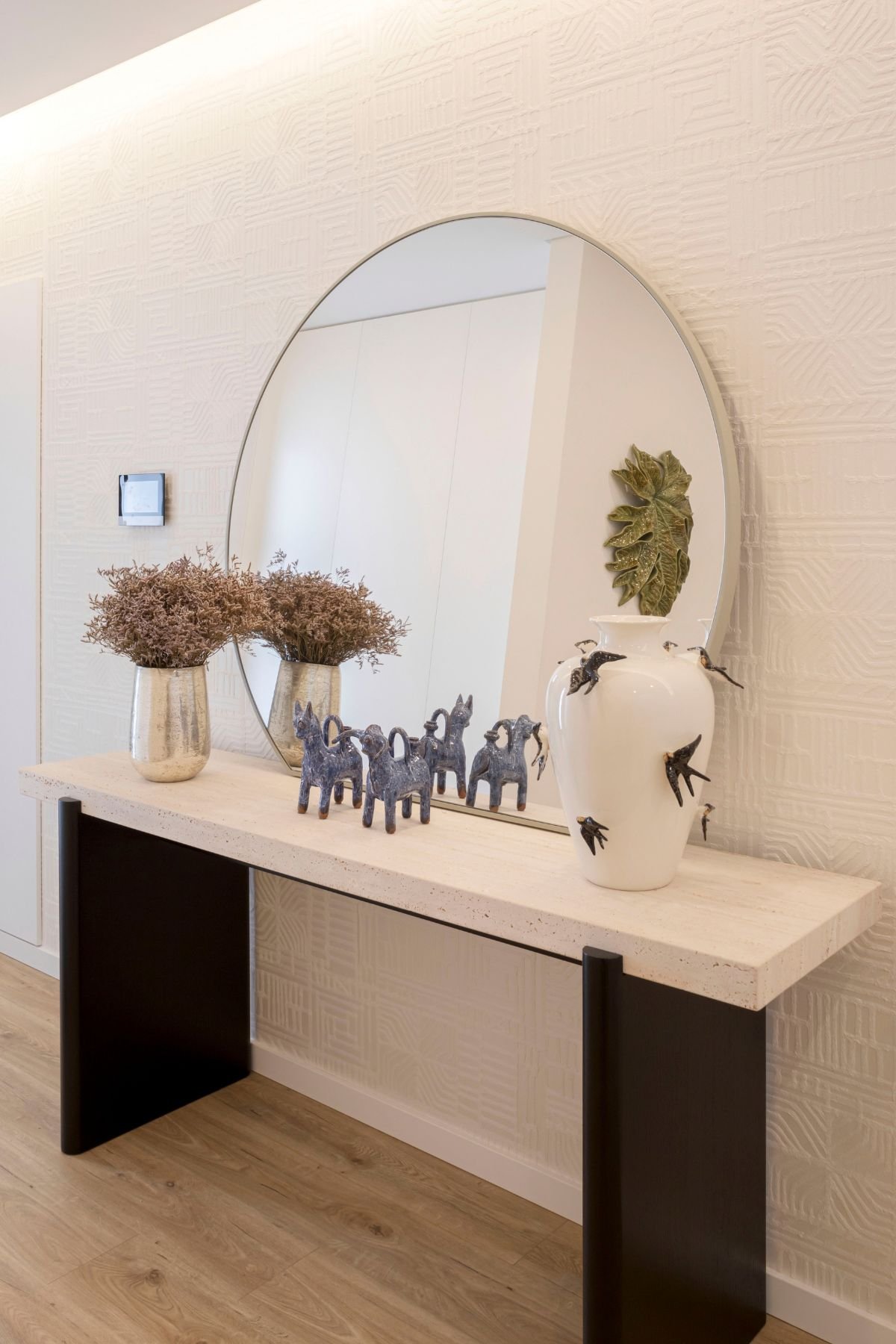 Enquadramento frontal do hall de entrada moderno, com a consola de entrada Natur decorada com peças Bordallo Pinheiro e um espelho grande Foz na parede com papel de parede textural.