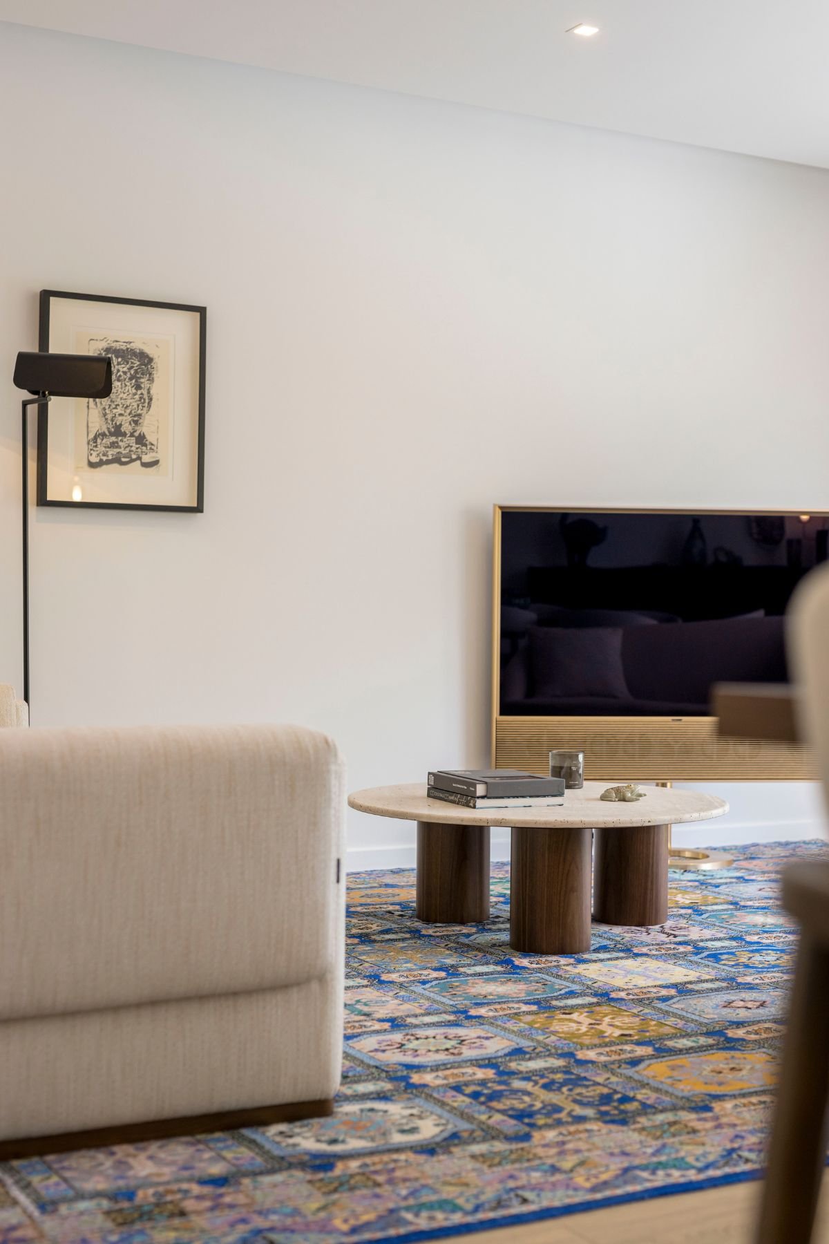 Detalhe da mesa de centro sala Natur em mármore travertino sobre tapete artesanal, com a televisão moderna ao fundo e quadros decorativos na parede.
