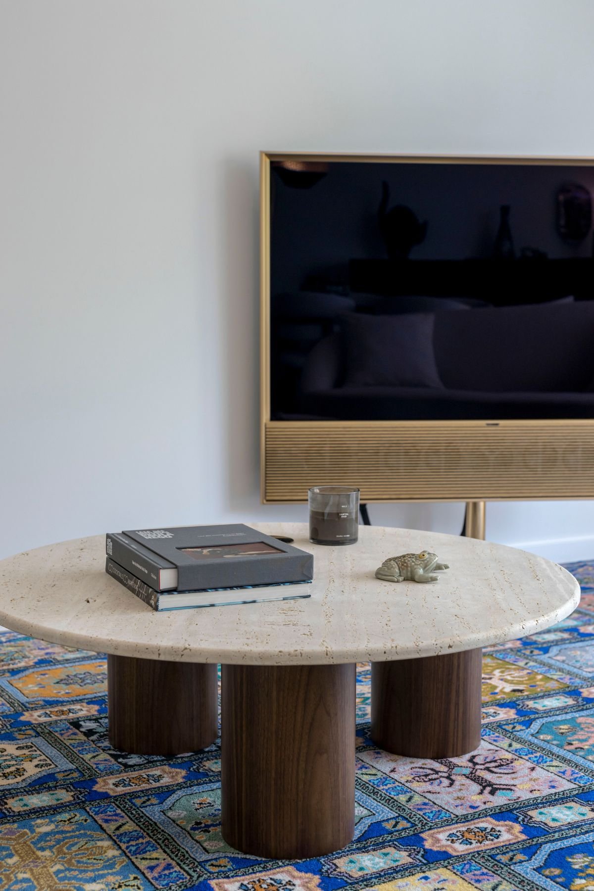 Pormenor da mesa de centro Natur em travertino com pés de madeira, velas aromáticas e livros decorativos sobre o tapete colorido na sala de estar moderna.