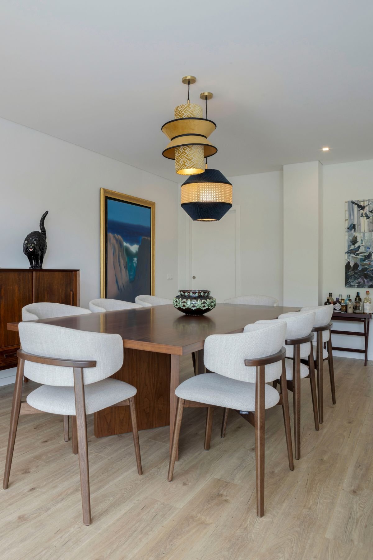 Ângulo da sala de jantar focando o conjunto de cadeiras de sala de jantar ao redor da mesa personalizada, com candeeiros suspensos e quadros decorativos ao fundo.