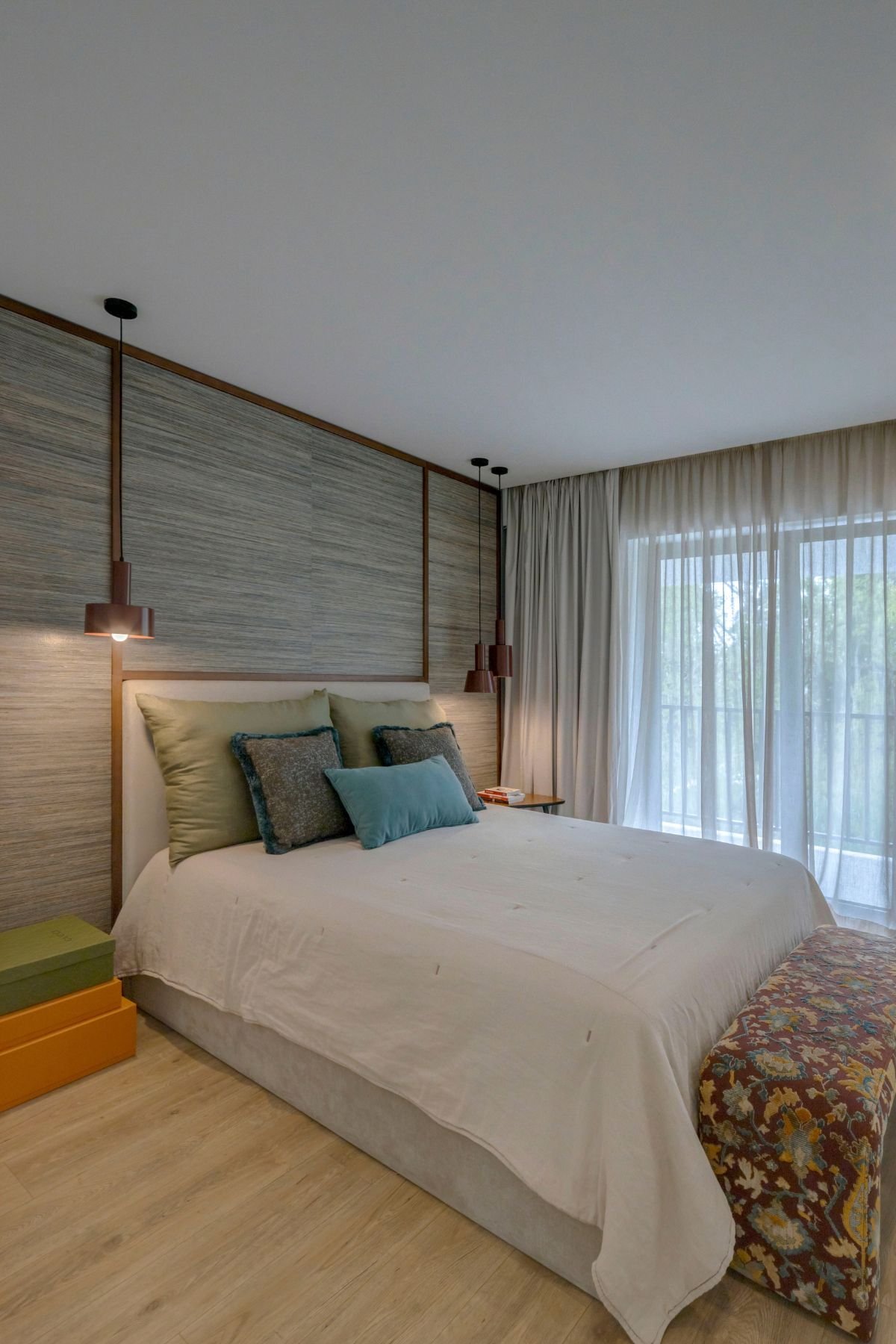 Perspetiva angular da cama de casal, mostrando o painel de parede, as iluminações modernas e o banco de apoio ao pé da cama estofado.