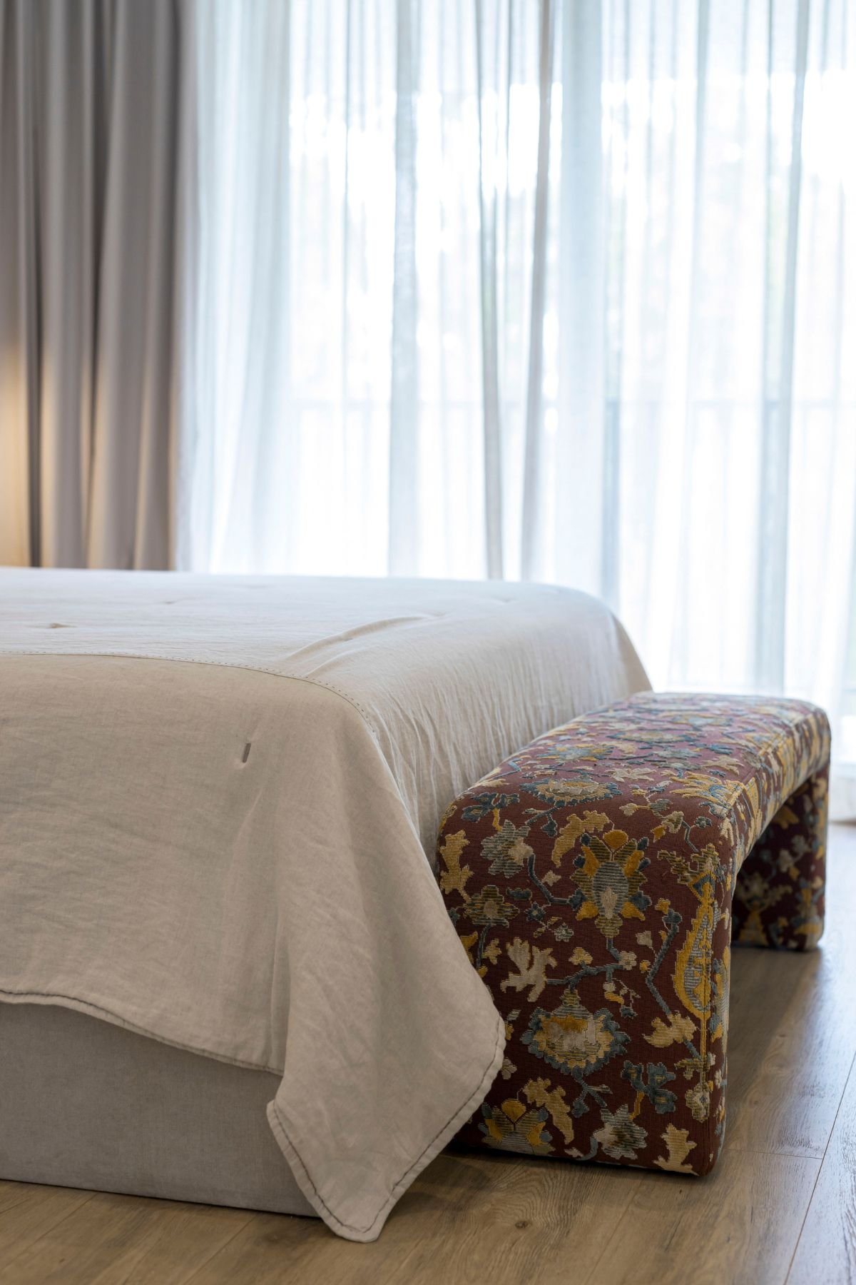 Pormenor do banco de apoio à cama estofado com padrão clássico/árabe, em contraste suave com os tecidos neutros do quarto.