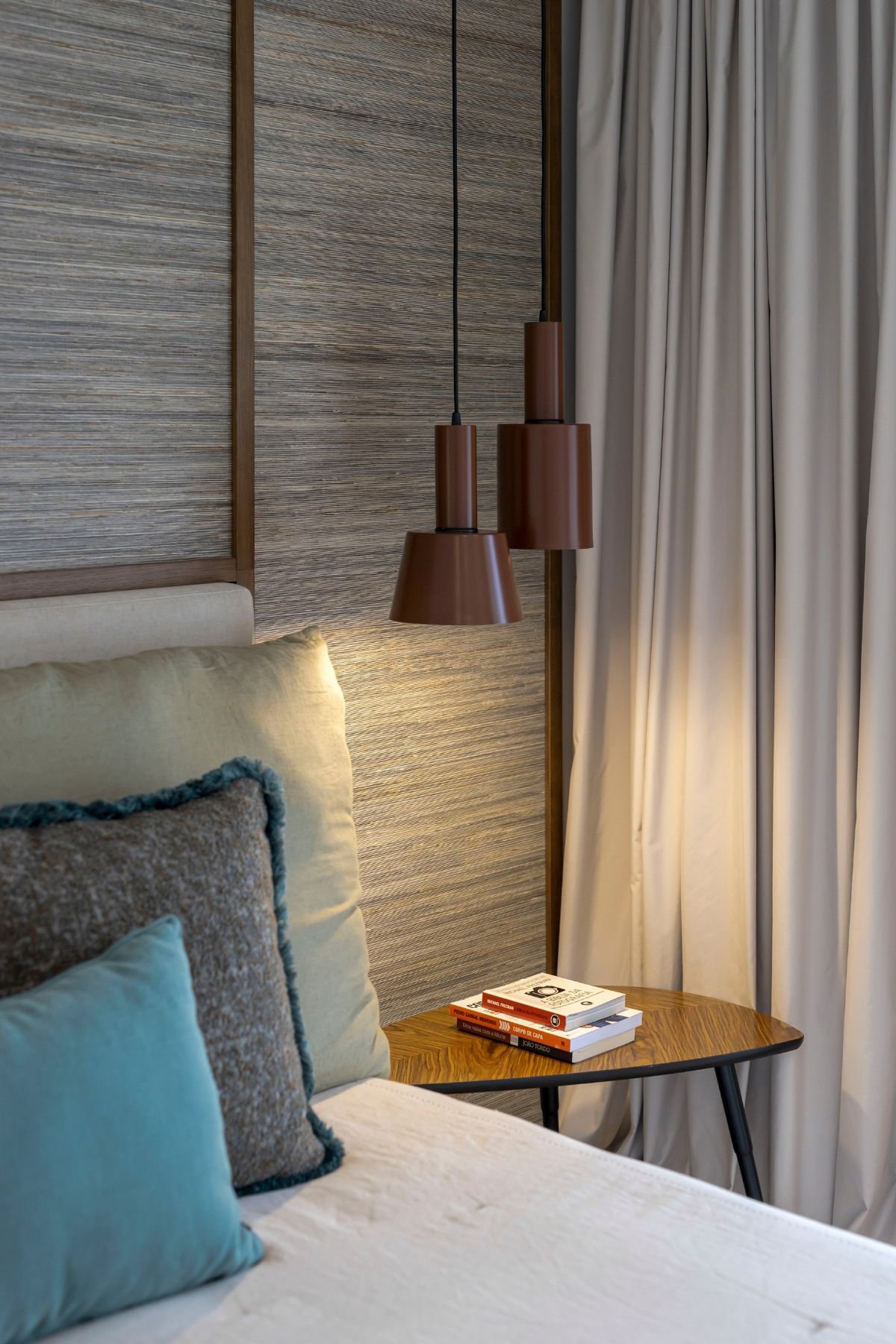Detalhe da mesa de apoio de cabeceira com livros, iluminação suspensa em tom terra e a textura acolhedora do painel de parede.