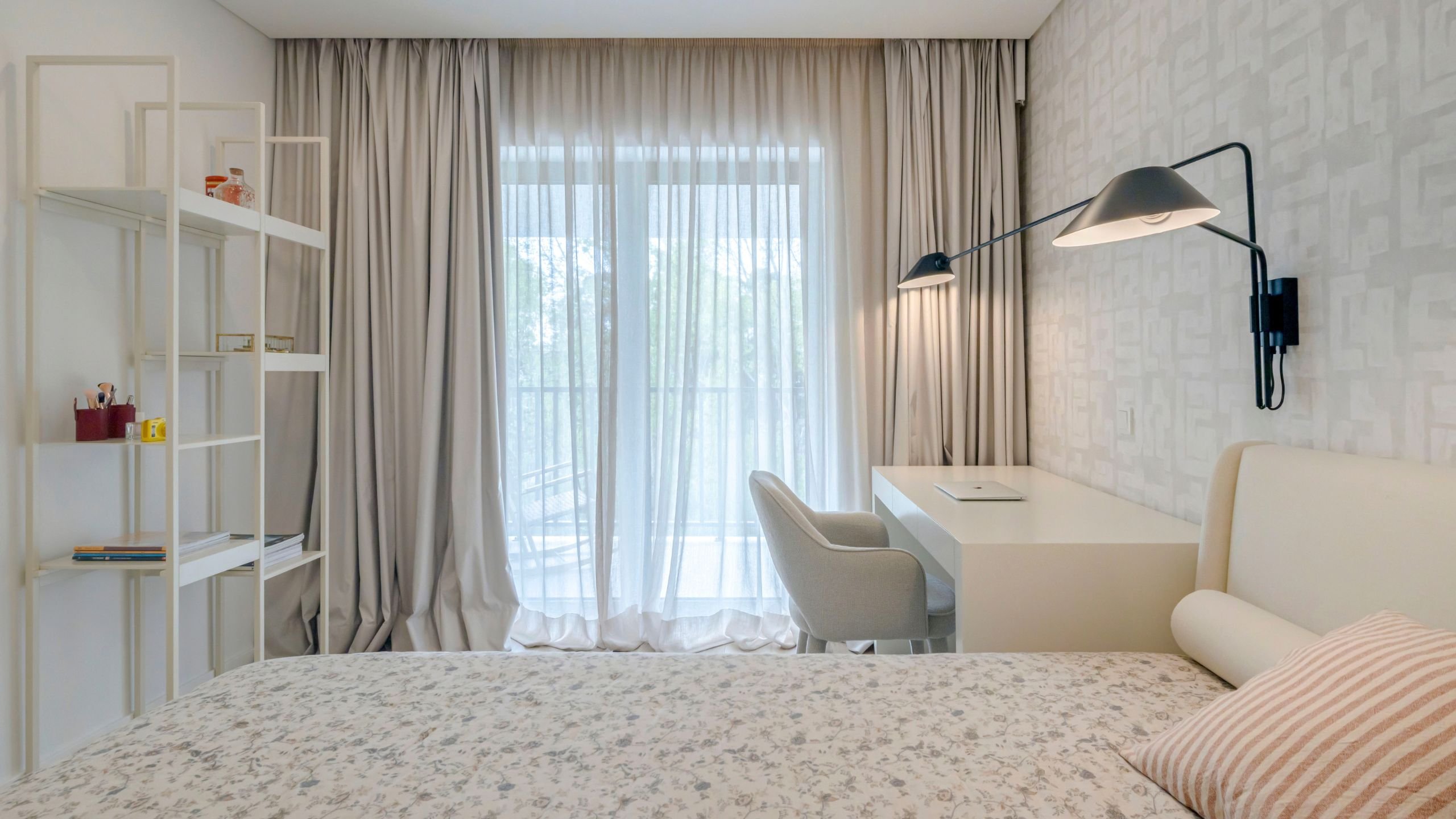 Visão geral da decoração de quarto, destacando a integração entre a cama com cabeceira estofada, a secretária branca com cadeira de apoio e a estante aberta de linhas minimalistas.