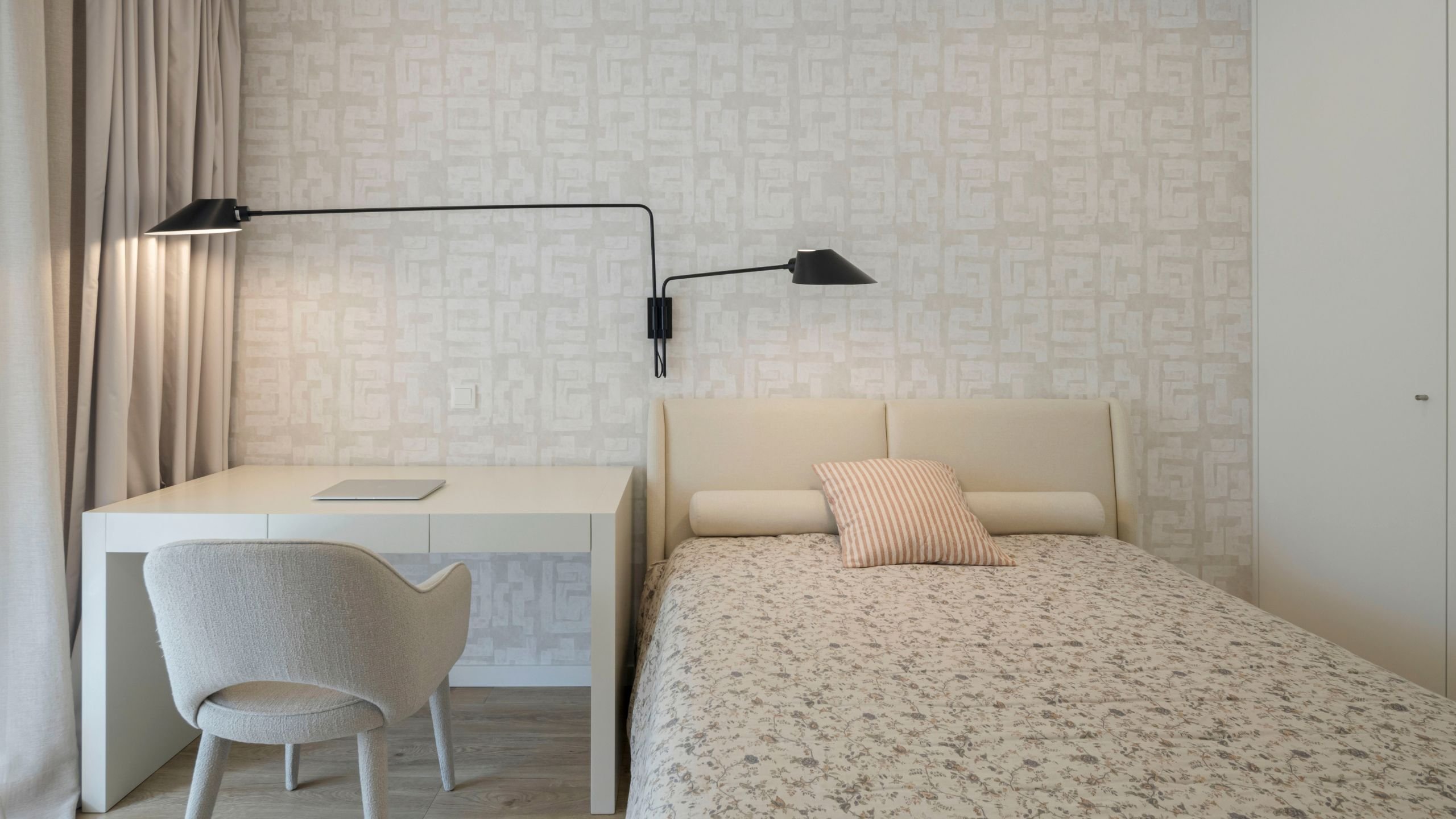 Vista frontal da cama e zona de trabalho, realçando a simetria do papel de parede texturado e o contraste dos apliques pretos de braço articulado sobre o tom claro do mobiliário.