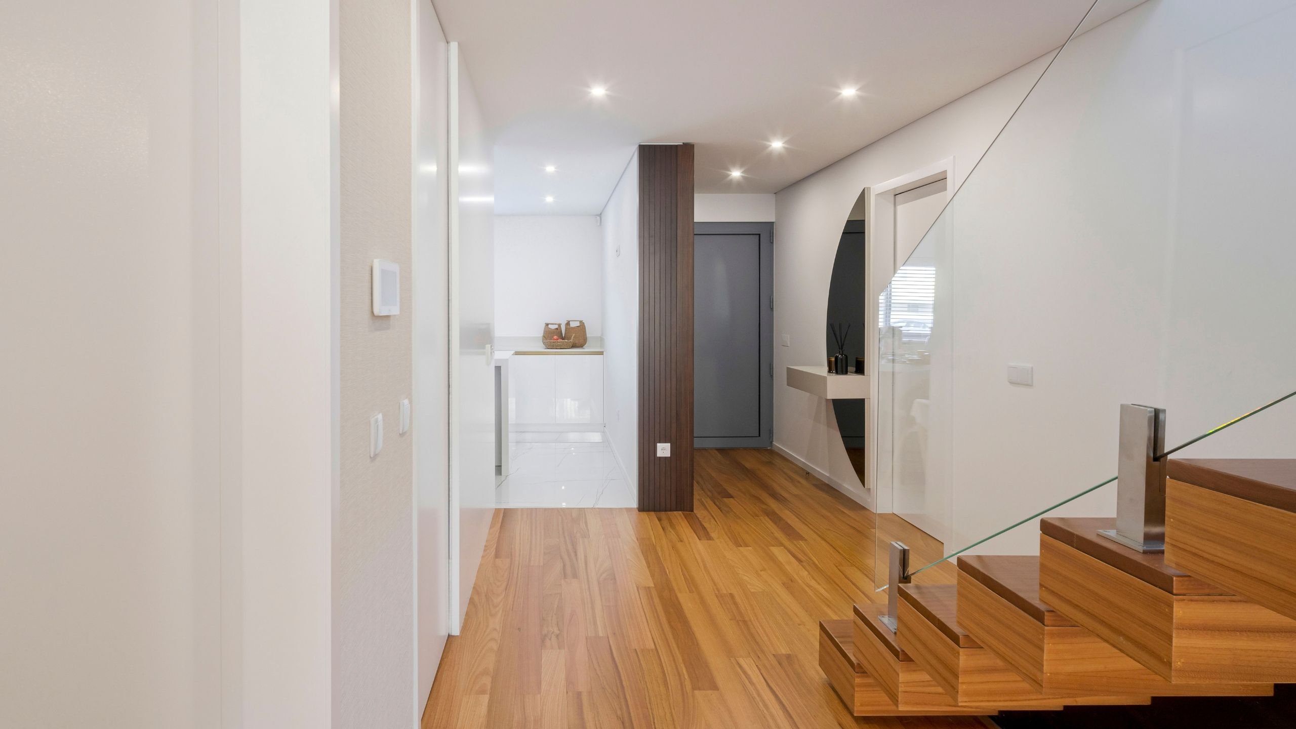 Hall de entrada moderno e discreto da Moradia Miramar com pavimento em madeira, pilar ripado escuro, escadas em vidro e consola suspensa com espelho meia-lua.