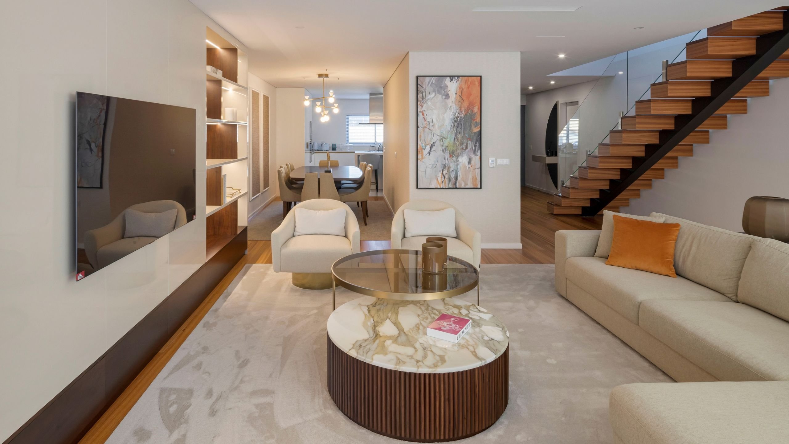 Visão geral da sala de estar moderna e luminosa na Moradia Miramar, destacando o sofá creme, as mesas de centro em mármore e nogueira, o móvel TV com estante e iluminação LED e a ligação fluida à zona de refeições.