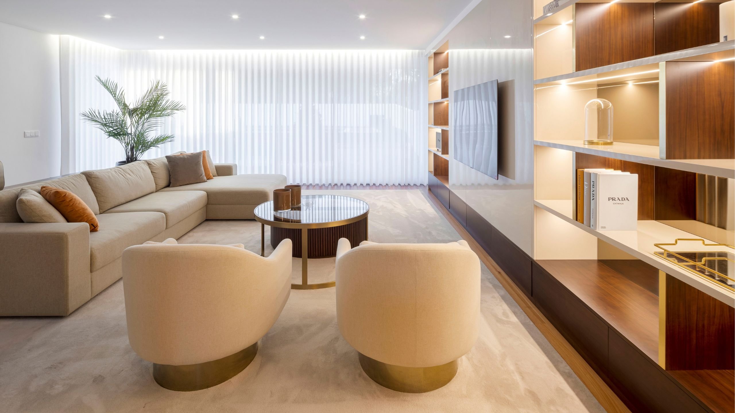 Perspetiva longitudinal da sala de estar focando nas duas poltronas creme com base metálica dourada e estante com luz LED.