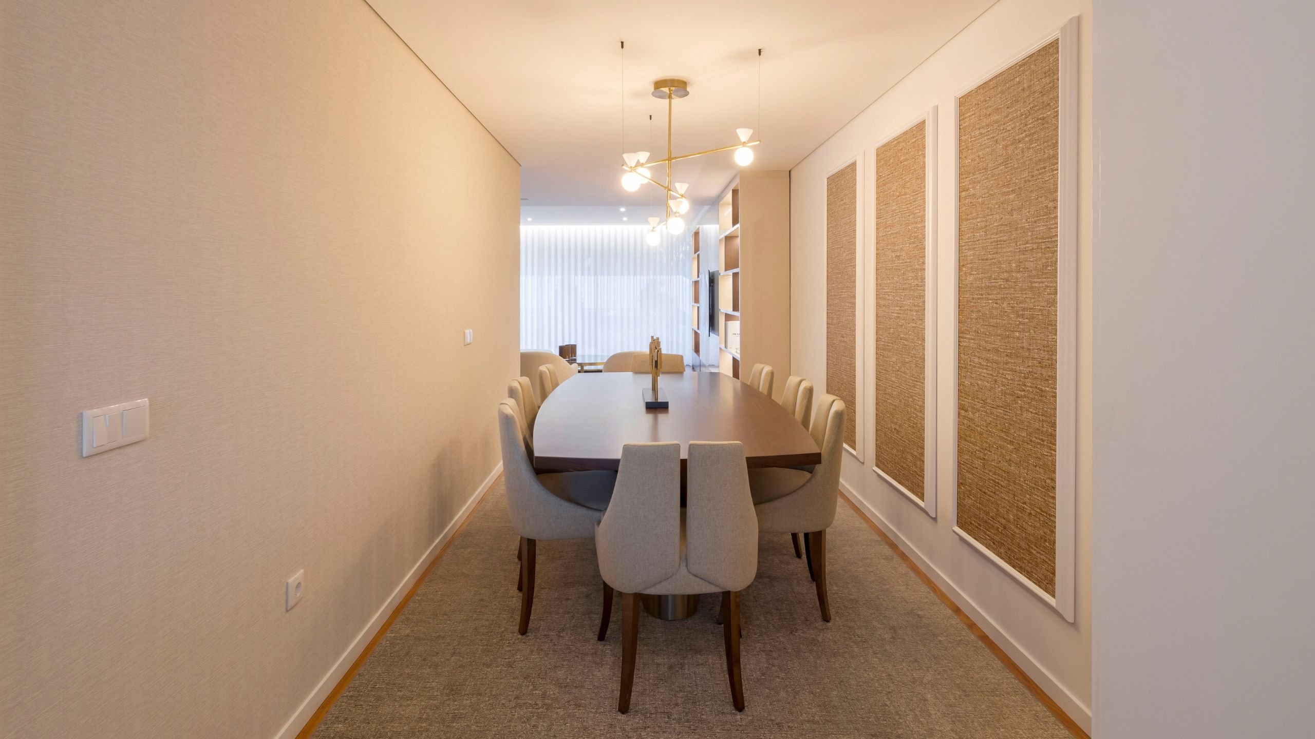 Visão geral da sala de jantar minimalista, destacando a mesa de jantar em nogueira, as cadeiras neutras e a estante de parede retroiluminada com decorações.