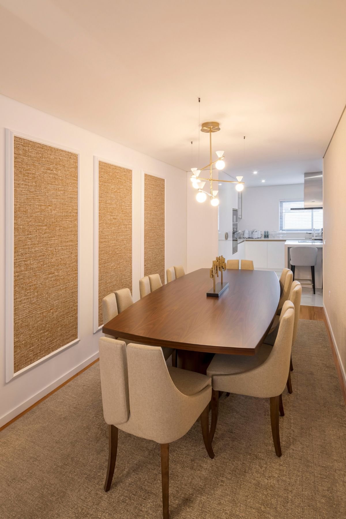Visão longitudinal da sala de jantar, com a mesa de nogueira alinhada com as cadeiras, sob o candeeiro suspenso e ladeada por painéis de parede texturizados.