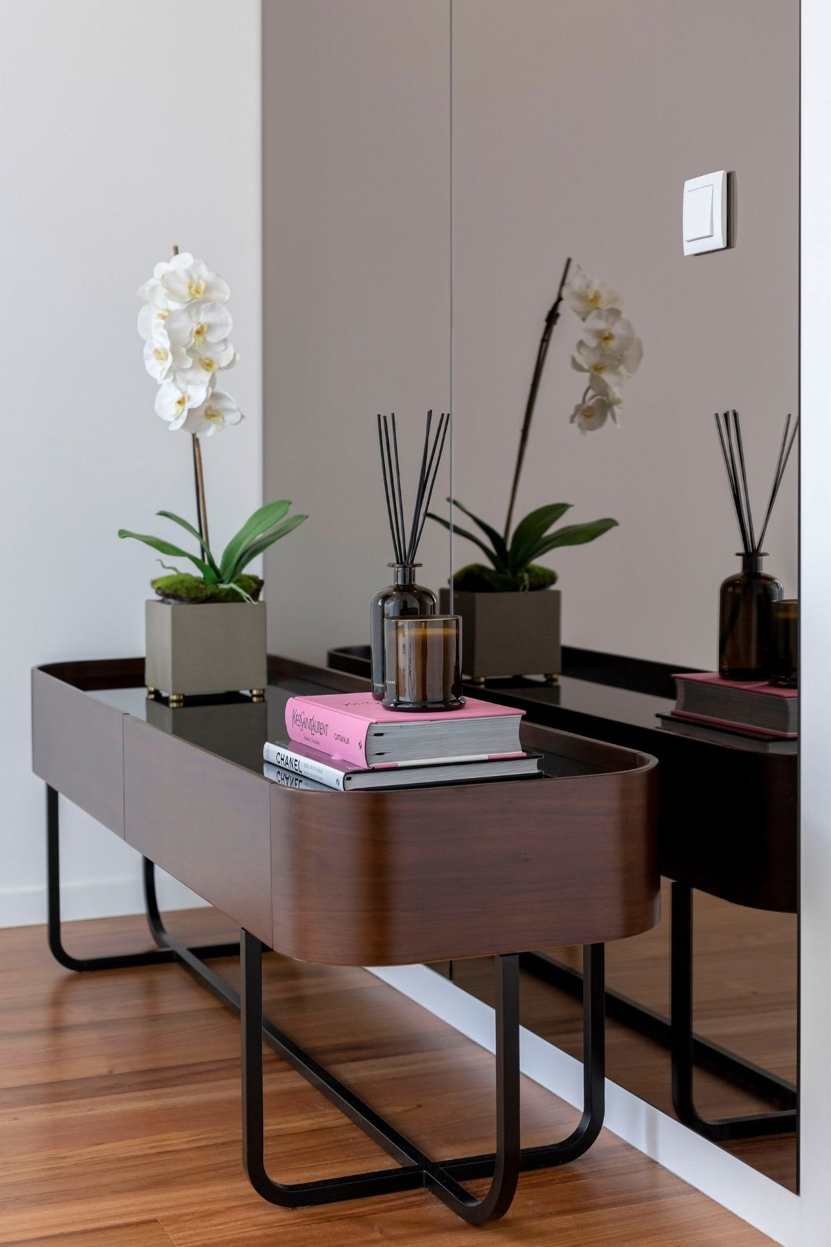 Pormenor da consola baixa em nogueira com estrutura em ferro mate, exibindo livros de design, fragrâncias e uma orquídea junto ao painel espelhado.