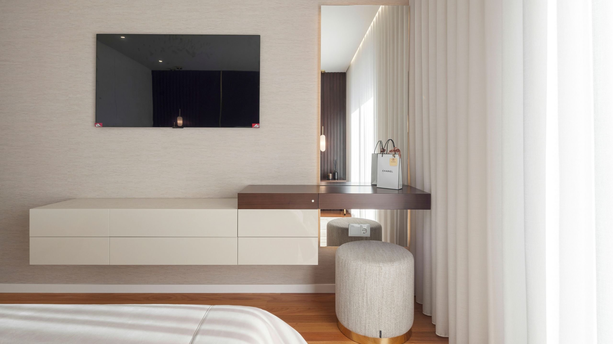 Visão geral da zona do toucador e móvel de TV do quarto suíte, destacando a consola suspensa neutra, o pufe estofado com base dourada e o espelho de corpo inteiro refletindo a luz natural.