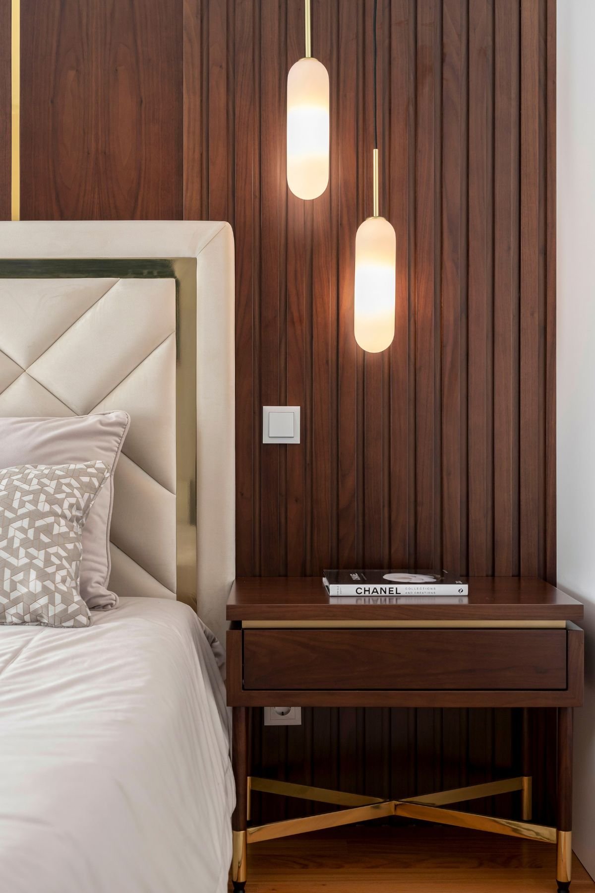 Pormenor da mesa de cabeceira em nogueira com base dourada, ao lado da cama de casal estofada, sob a luz suspensa vertical de design contemporâneo.