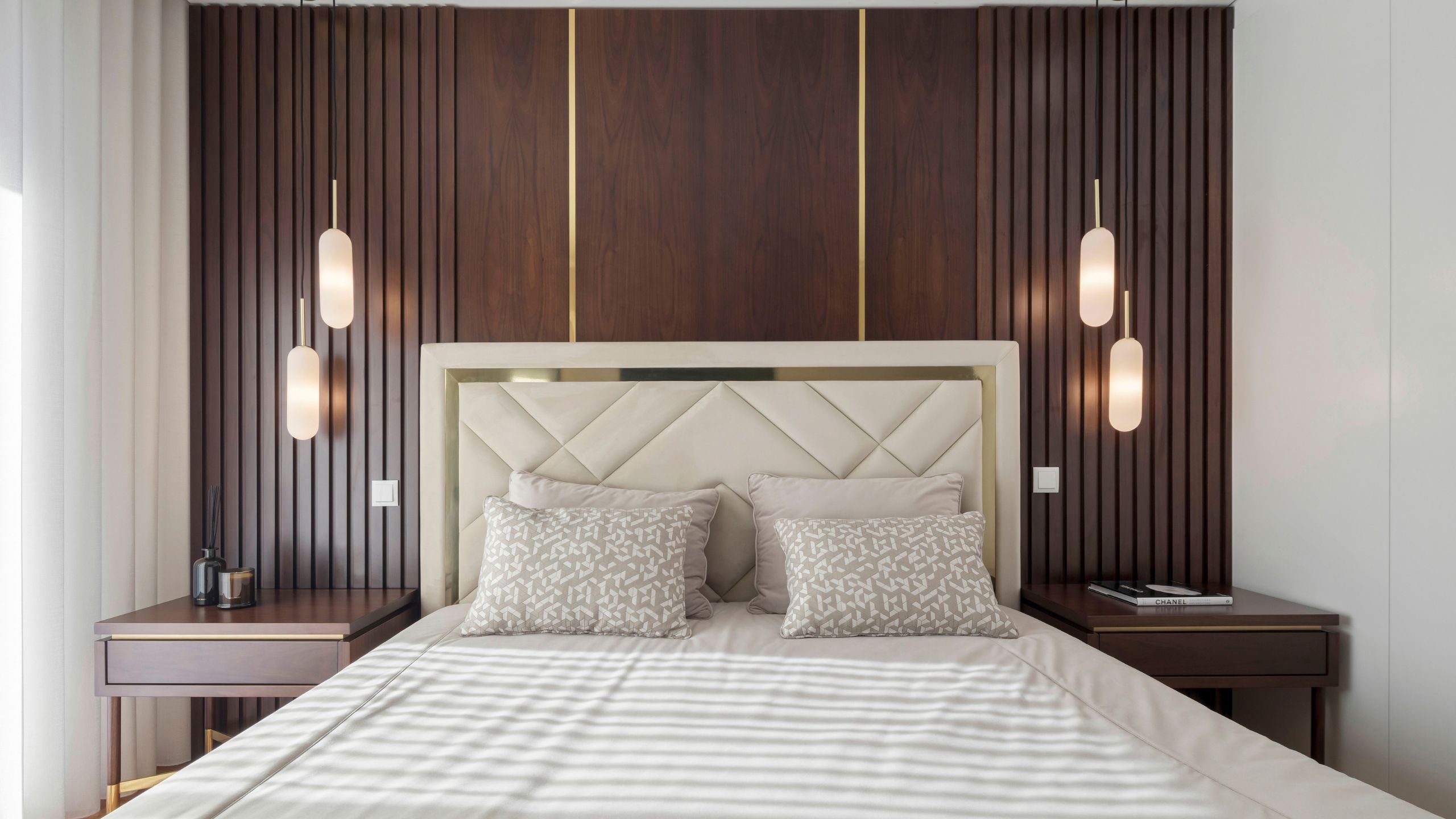 Vista frontal da cama de casal e mesas de cabeceira, realçando a simetria do painel ripado de nogueira com detalhes em latão e os pendentes luminosos verticais.