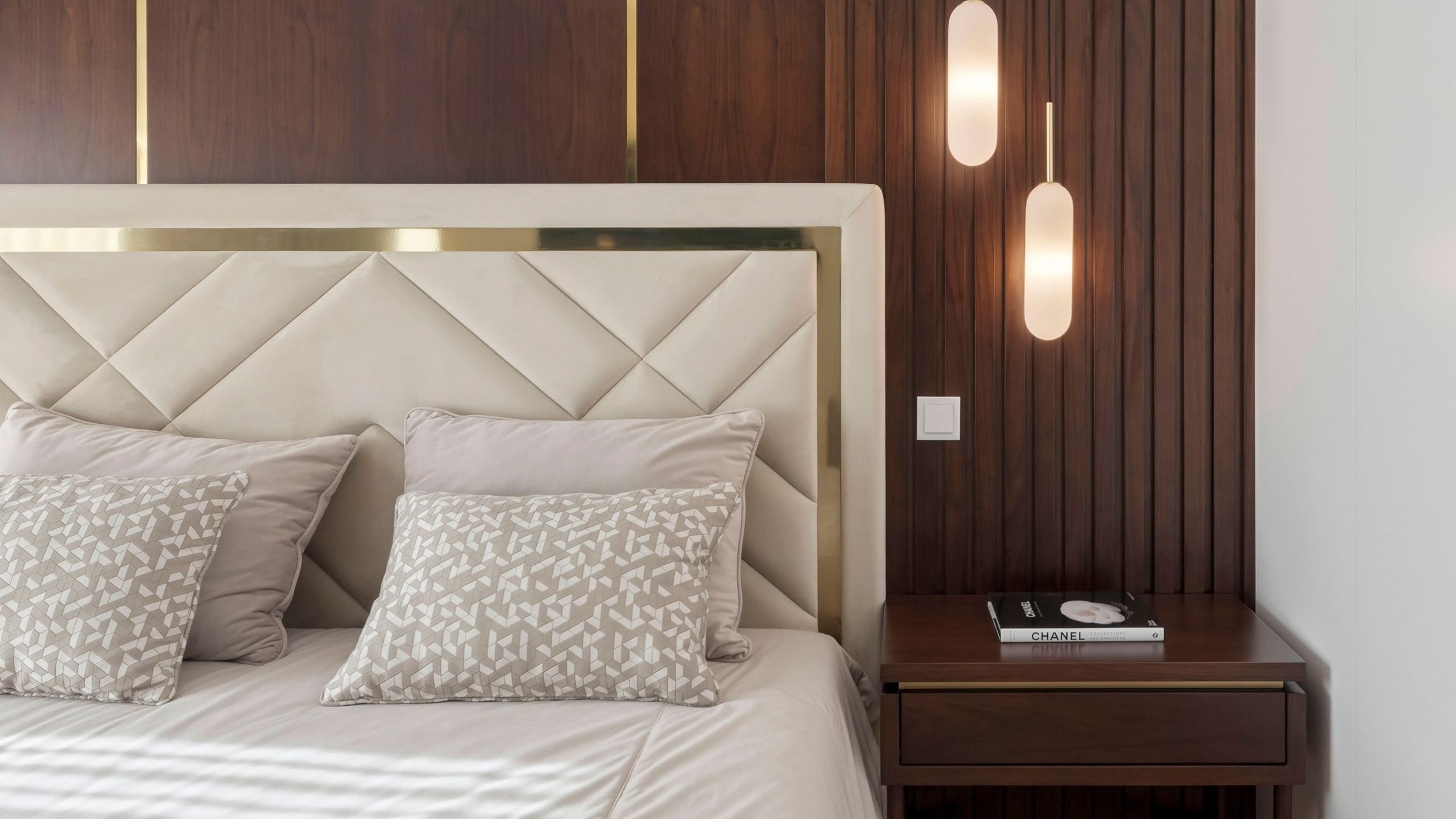 Plano aproximado da cabeceira de cama estofada com padrão geométrico e moldura dourada, ao lado da mesa de cabeceira com livro de design.