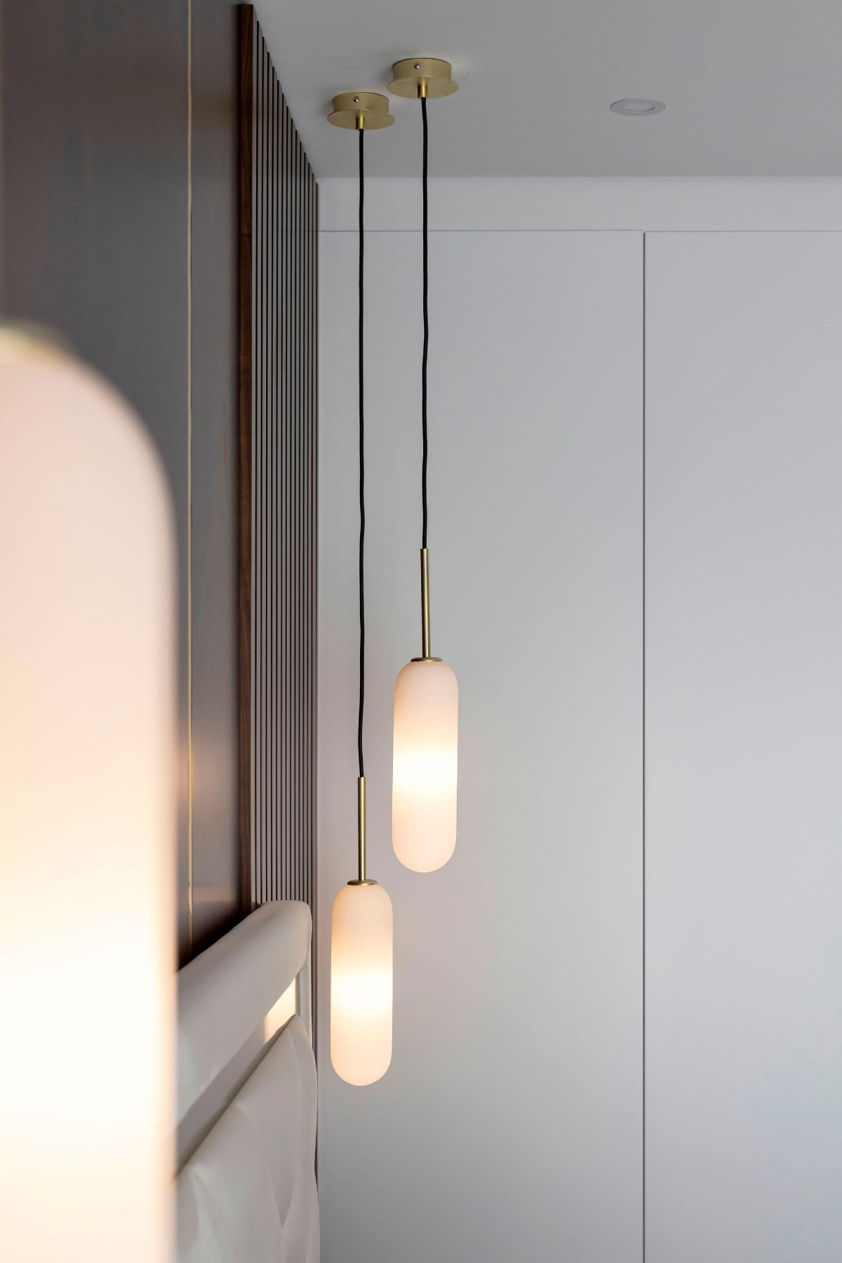 Detalhe aproximado dos pendentes luminosos verticais com acabamento em latão, suspensos sobre a mesa de cabeceira, junto ao painel ripado de nogueira.