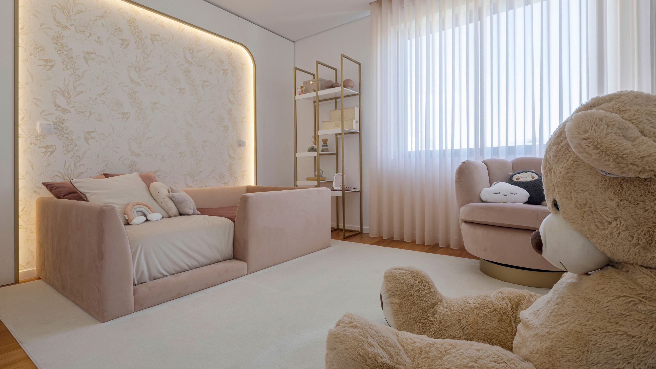 Perspetiva angular do quarto de princesa, mostrando a cama de criança rosa aveludada, o painel de parede curvo e retroiluminado com papel de parede dourado e o conjunto de estantes branco e dourado.