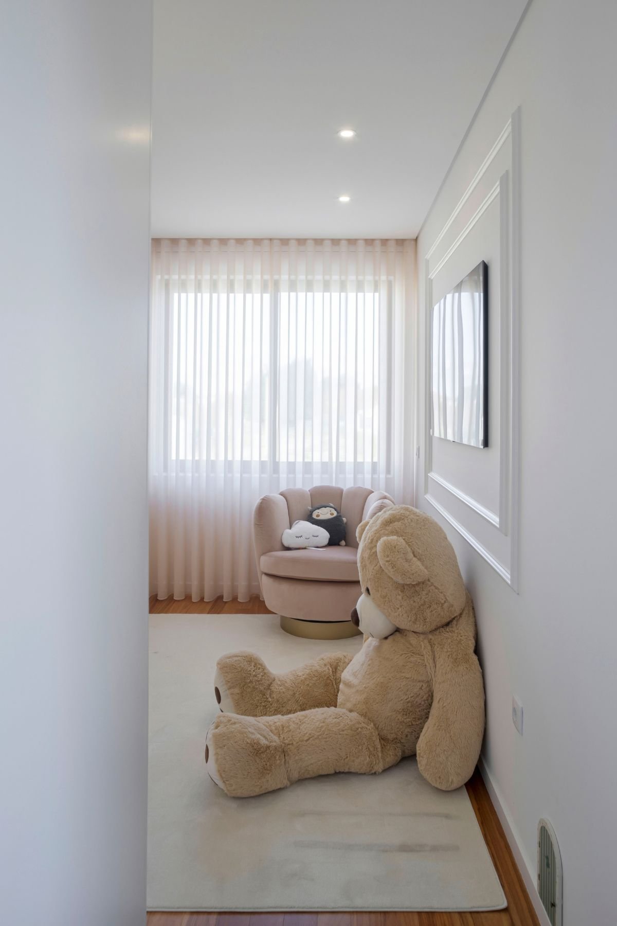 Visão geral do corredor de entrada para o quarto de criança, destacando a cortina rosa translúcida, o cadeirão rotativo estofado em veludo rosa com base dourada e um peluche grande sentado no chão.