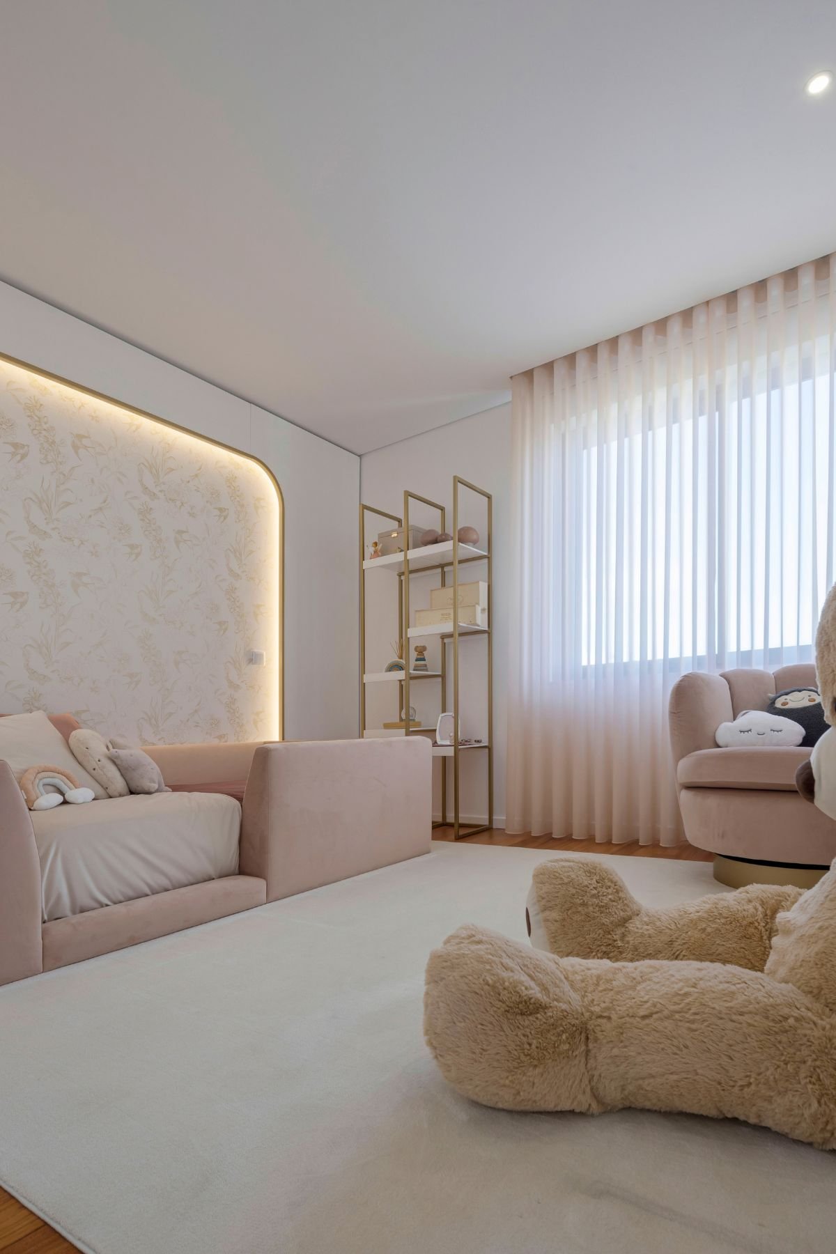 Perspetiva angular do quarto de princesa, mostrando a cama de criança rosa aveludada, o painel de parede curvo e retroiluminado com papel de parede dourado e o conjunto de estantes branco e dourado.