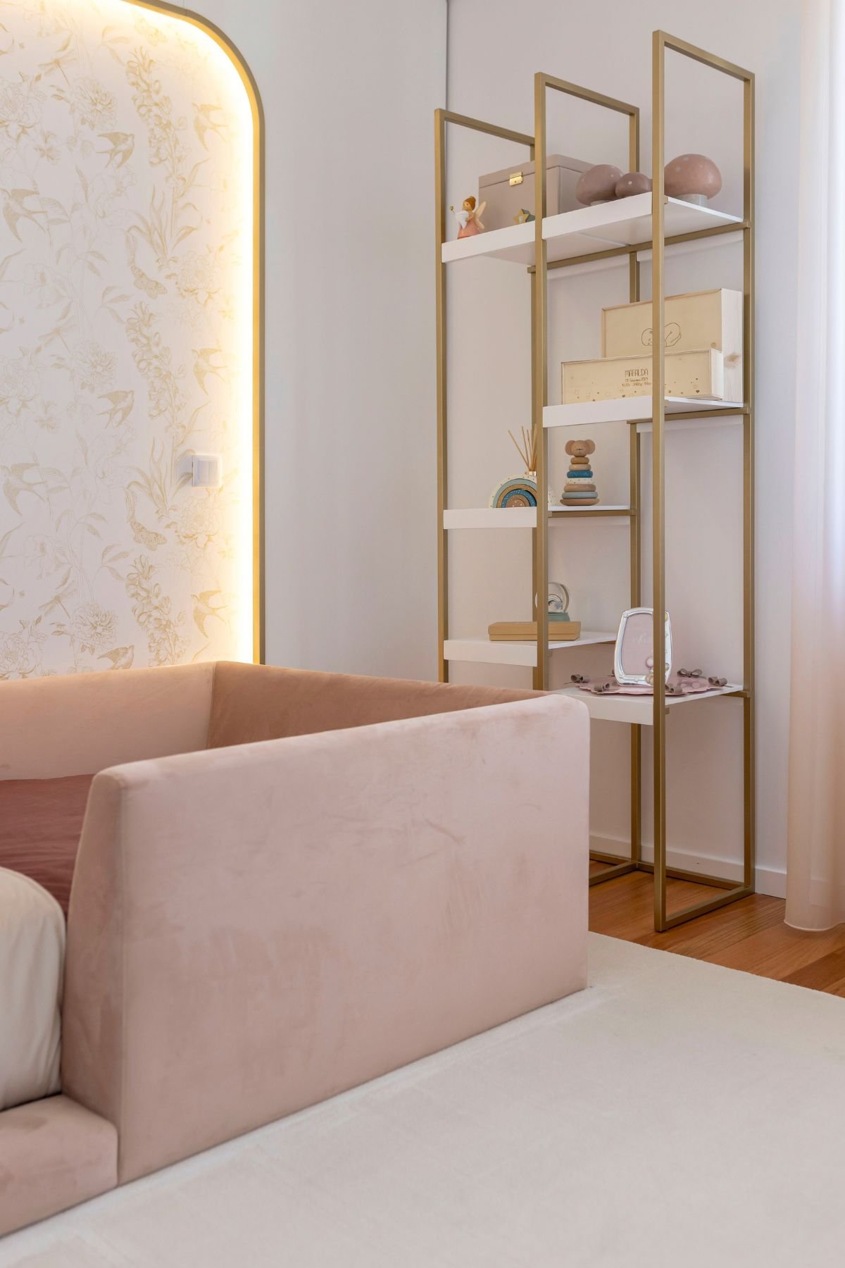 Visão de detalhe do quarto de criança, focando no conjunto de estantes dourado e branco decorado com brinquedos e acessórios infantis, ao lado da cama rosa aveludada.