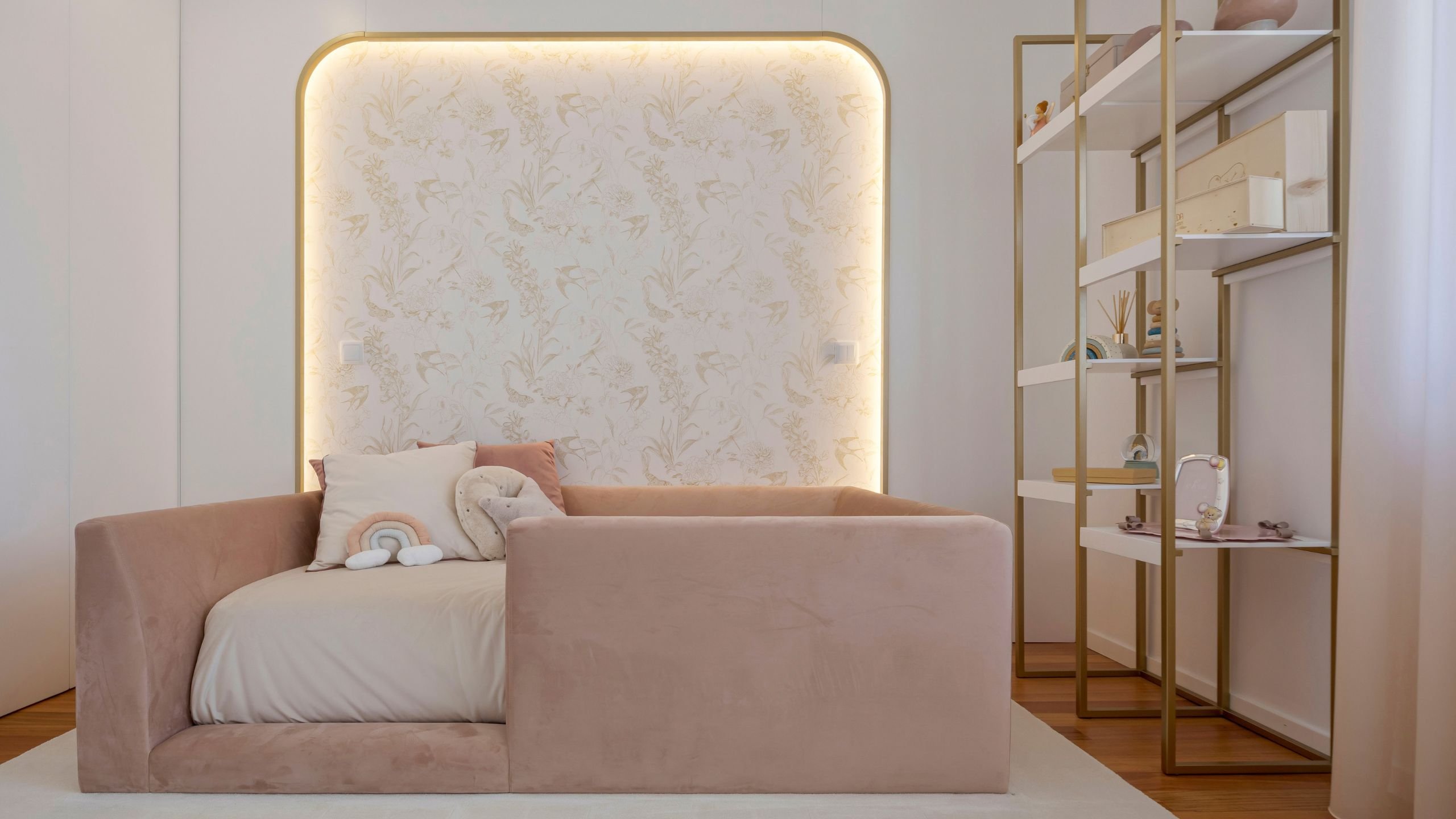 Visão frontal enquadrada da cama de criança rosa no quarto de princesa, posicionada centralmente em frente ao painel de parede com papel de parede floral dourado e iluminação suave.