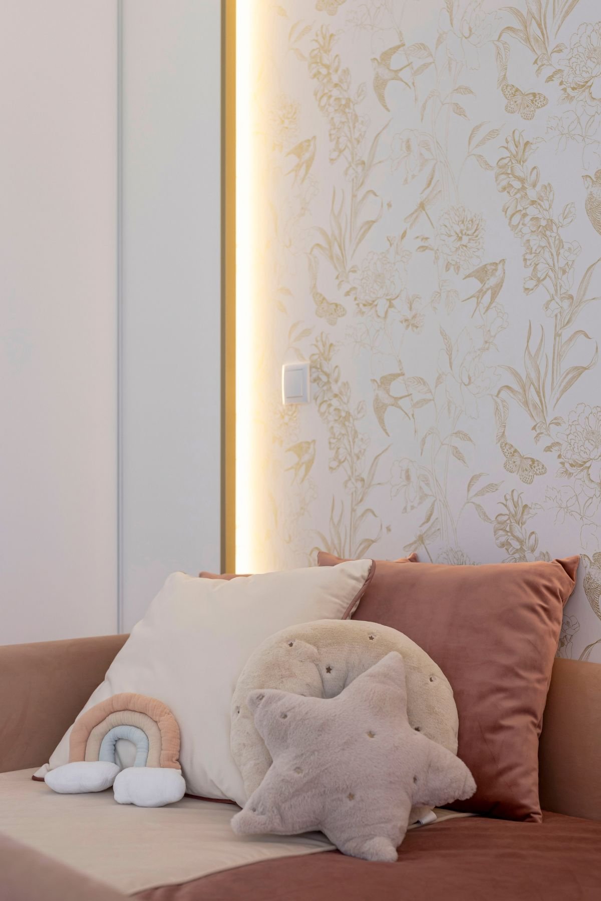 Pormenor lateral da cama de criança no quarto de princesa, realçando a textura do estofo rosa e o contraste com o papel de parede dourado e a luz quente embutida.