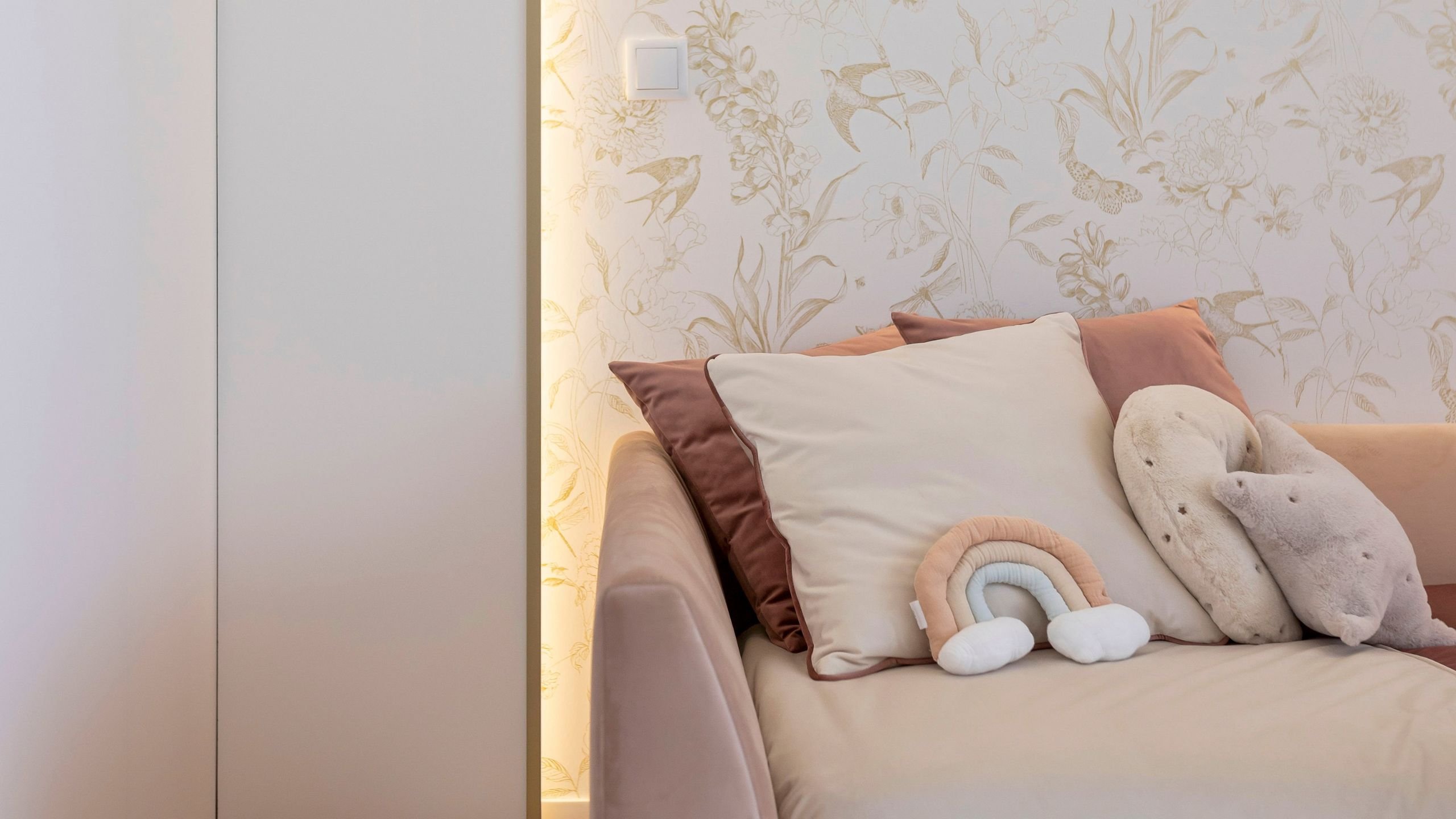 Detalhe aproximado das almofadas e peluches sobre a cama de criança no quarto de princesa, com foco na textura dos tecidos e no papel de parede dourado retroiluminado ao fundo.