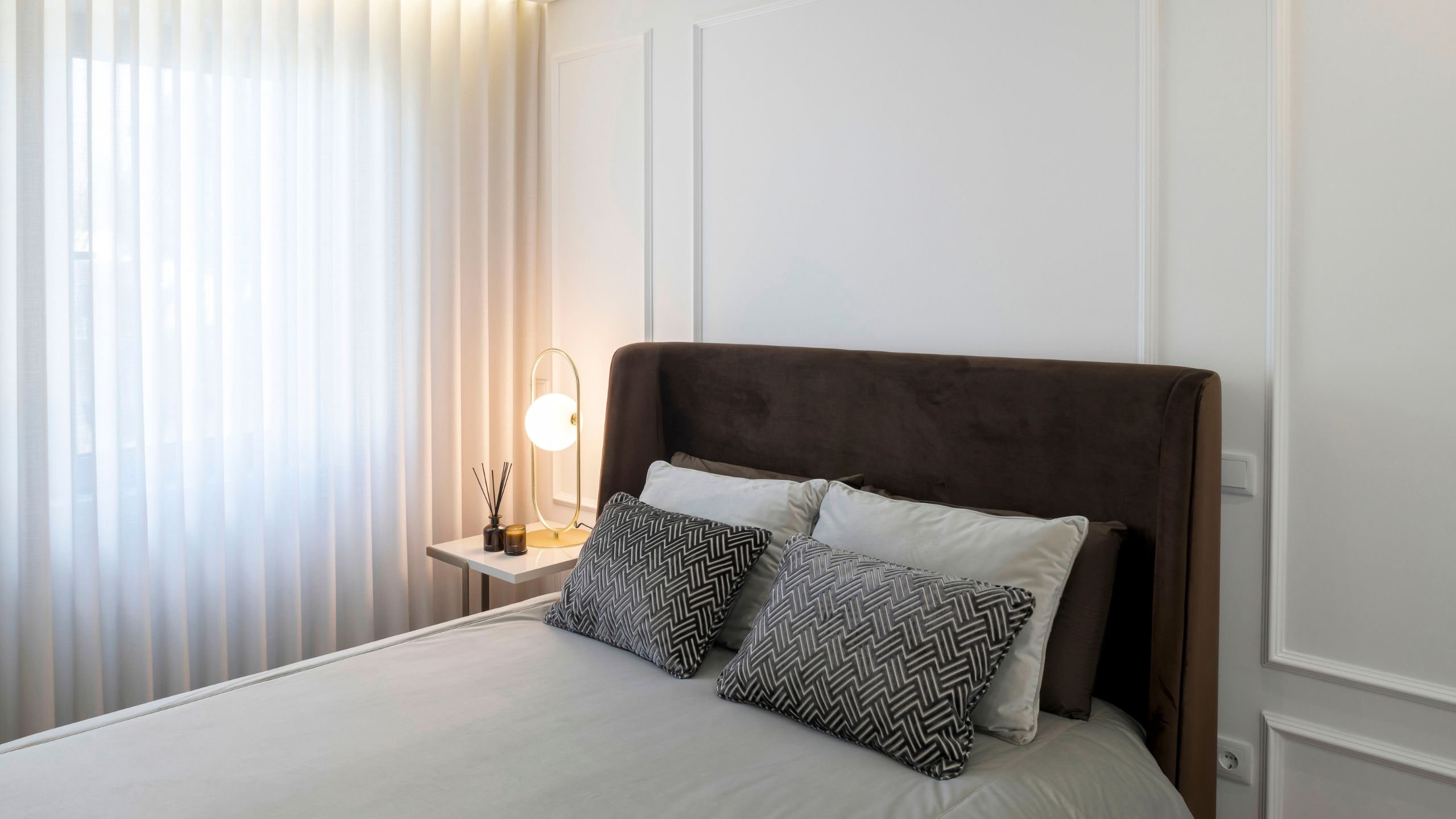 Visão geral do quarto de hóspedes, com foco na cama de cabeceira estofada em tom castanho escuro, na mesa de apoio ao lado da janela e no trabalho de molduras clássicas nas paredes.
