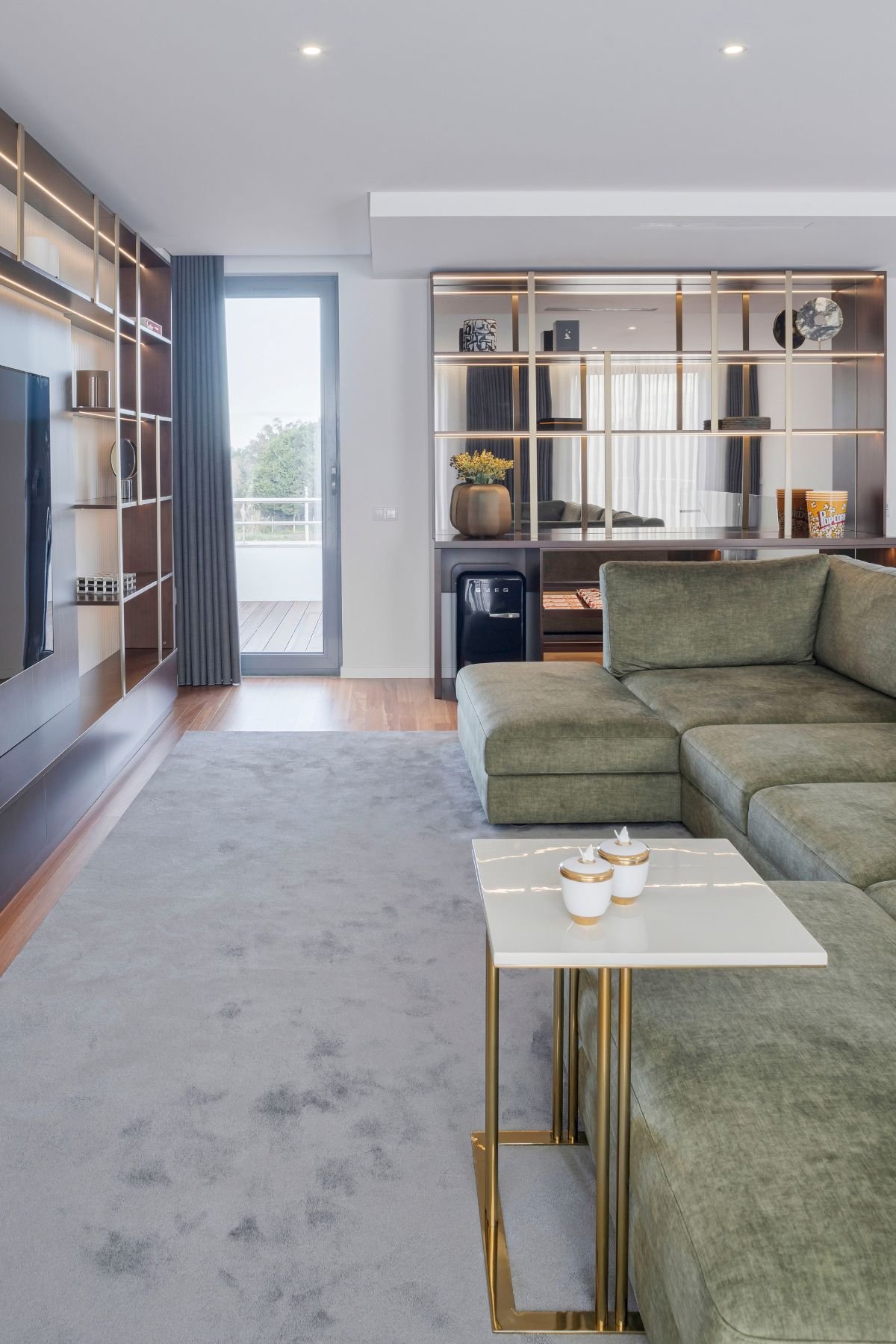 Visão geral do home cinema contemporâneo, realçando o sofá de canto em tom verde aveludado, o tapete cinzento e o móvel bar em nogueira e latão ao fundo. Em primeiro plano, a mesa de apoio com tampo bege brilhante e dois potes decorativos.