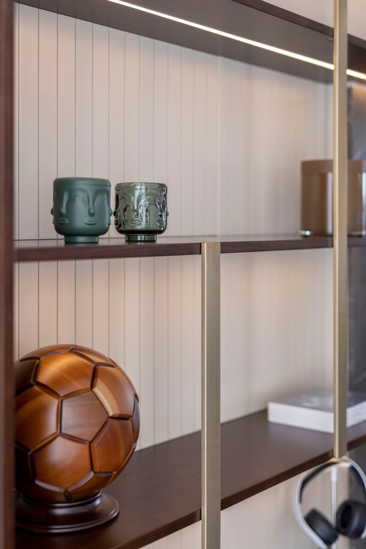 Pormenor focado nas prateleiras da estante, realçando os acabamentos em latão mate e a iluminação LED embutida que destaca os vasos decorativos e a bola de futebol.