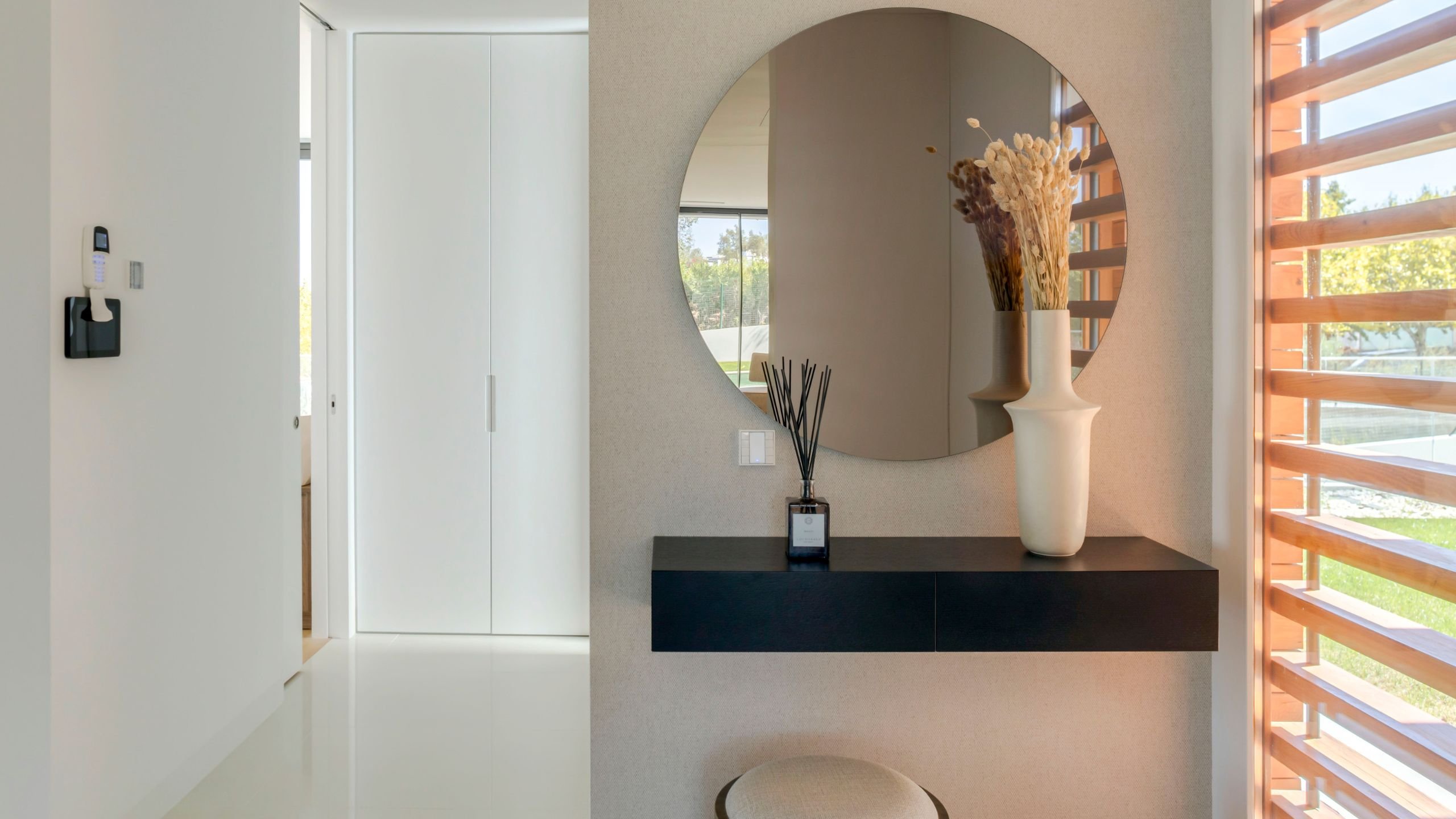 Entrada da Moradia Oeiras com consola suspensa preta minimalista, espelho redondo, vaso decorativo com espigas secas e ambientador de varetas.