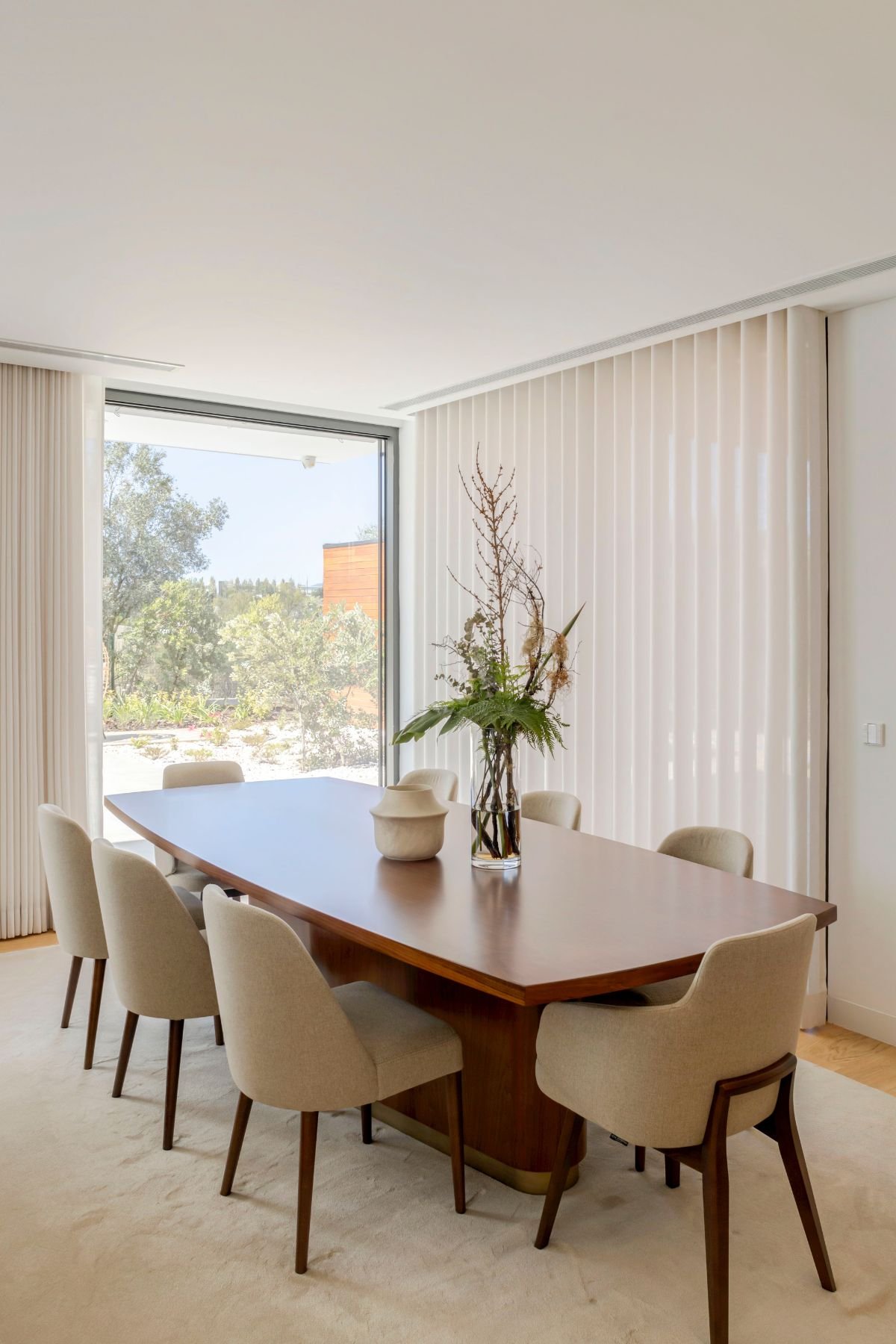 Mesa de jantar em madeira com pé de detalhe em latão escovado, rodeada pelas cadeiras em tom creme e iluminação natural de fundo.