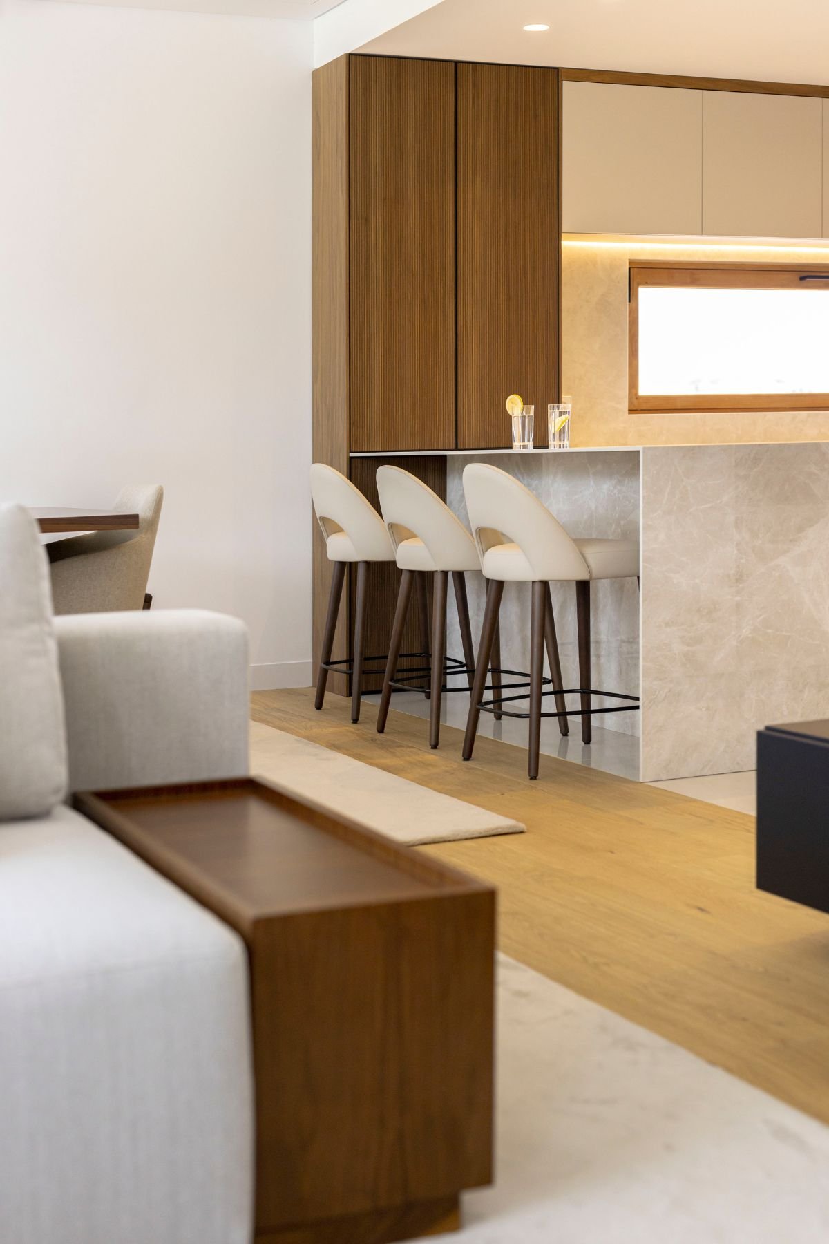 Perspetiva lateral da bancada da cozinha com cadeiras altas em tom creme e pés escuros, em plena harmonia com a sala.