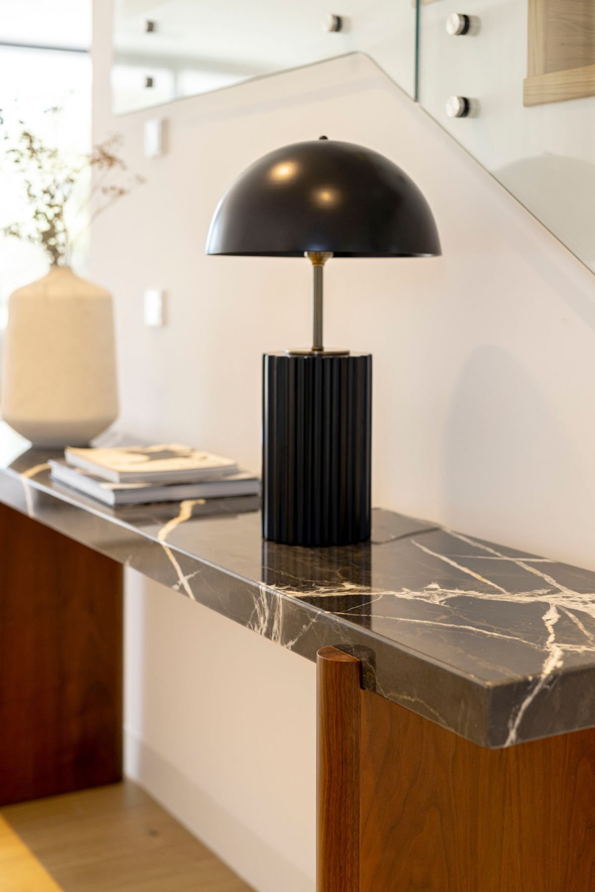 Pormenor da consola com tampo em mármore escuro com veios brancos, pés em madeira, candeeiro de mesa preto de design moderno e vaso cerâmico branco.