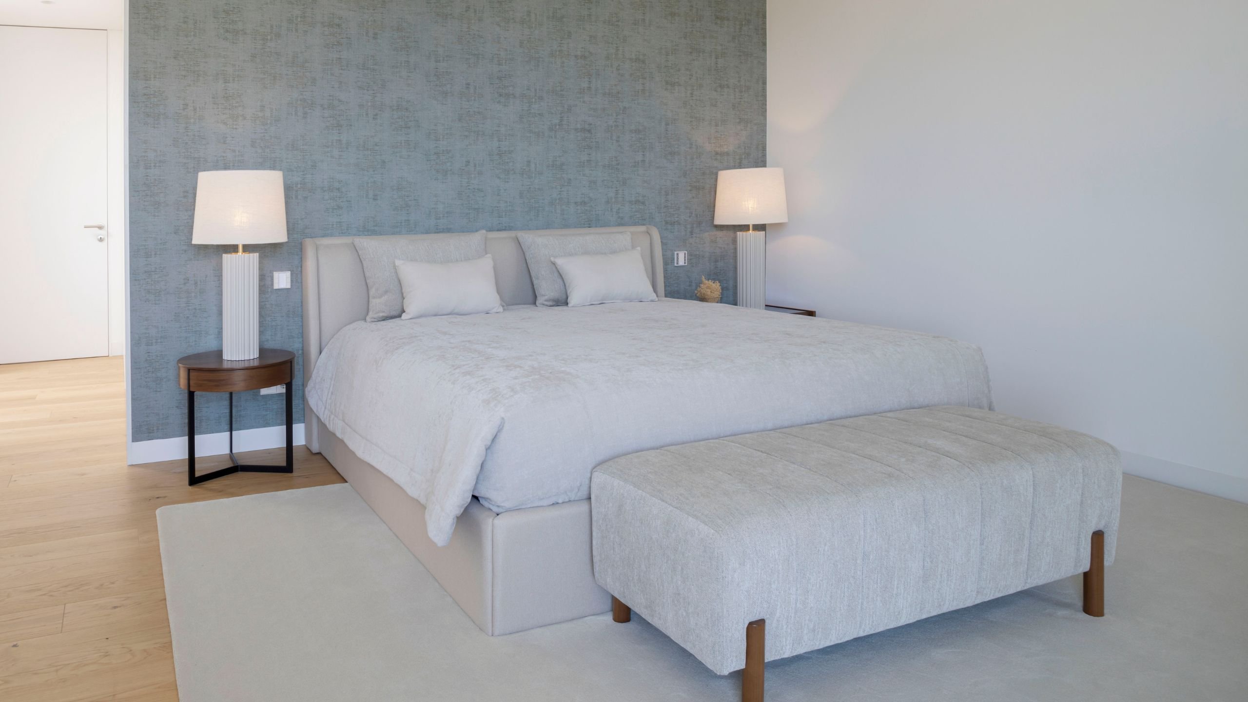 Visão frontal da cama com cabeceira curva em tecido claro, banqueta aos pés, papel de parede azul e mesas de cabeceira em madeira escura com candeeiros de mesa.